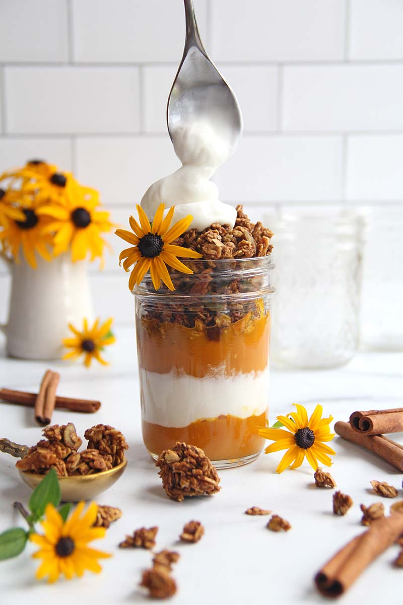 Spoon adding a dollop ofyogurt on top of a pumpkin breakfast parfait with layers of sweetened and spiced pumpkin puree, yogurt, and pumpkin granola.
