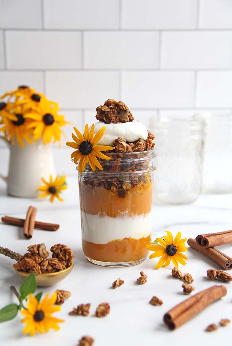 Pumpkin yogurt parfait, shown in a mason jar in a white kitchen, next to cinnamon sticks and a small bowl of pumpkin granola.