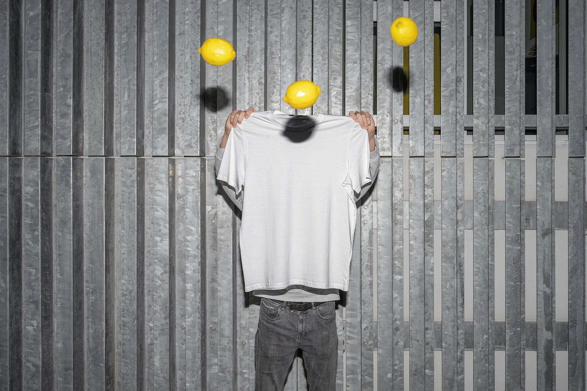 Blank T-Shirt Mockup held in front of a metal wall with floating lemons and soft shadows.