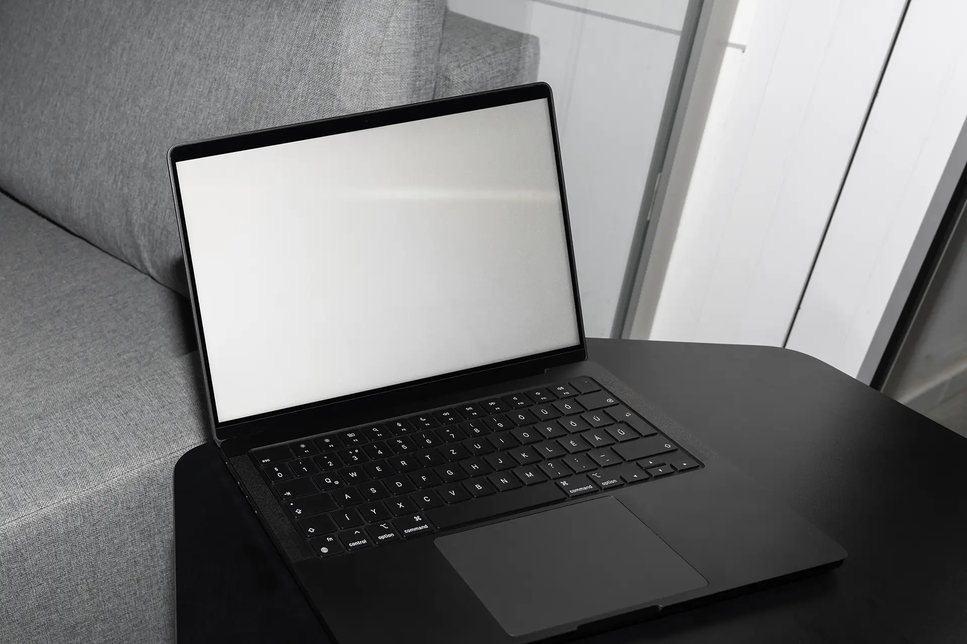 Blank MacBook Mockup with empty screen on black table next to grey sofa.