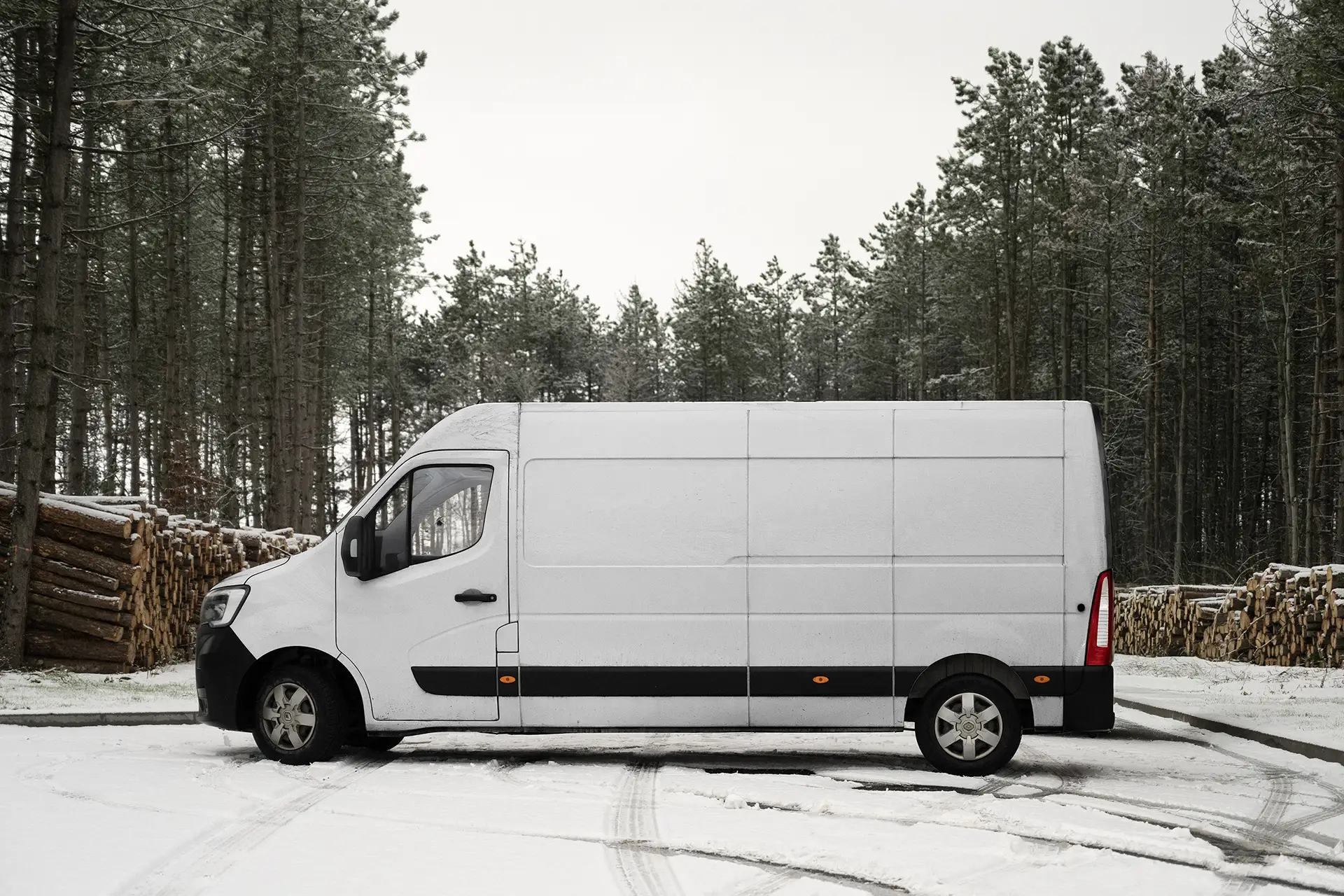 Blank white delivery van mockup photographed in a snowy forest setting with stacked logs and soft diffused overcast light.