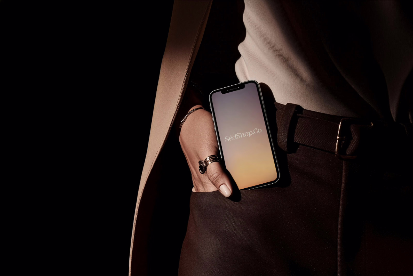An iPhone held at the hip showing a warm gradient SedShop.Co screen, in a dark, tailored, cinematic setting.