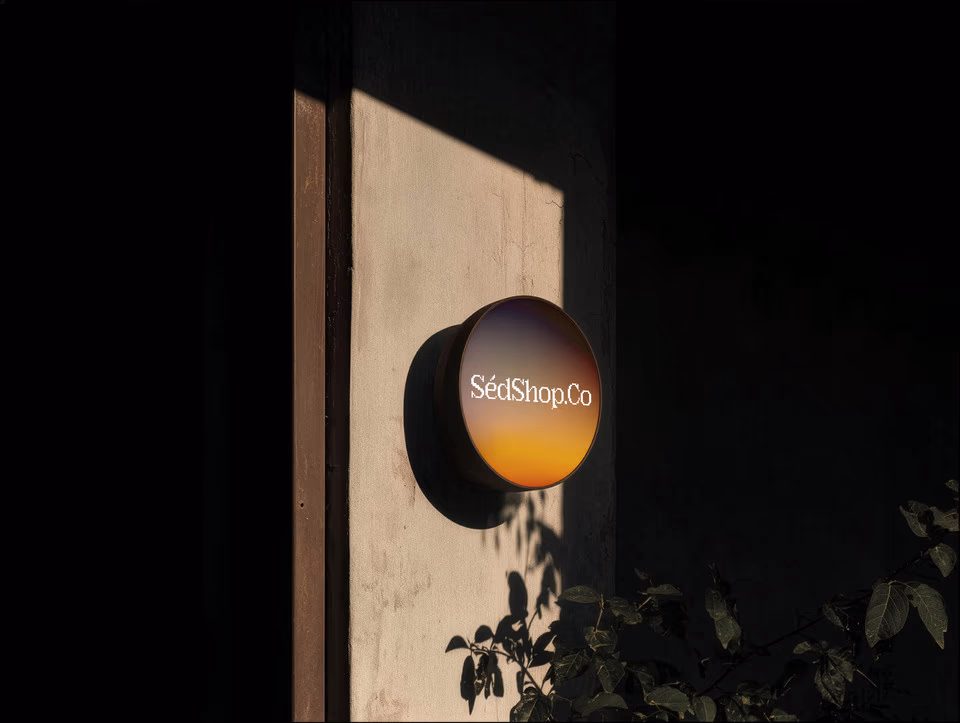 A round illuminated lightbox sign with a warm gradient design on a dark exterior wall with foliage.