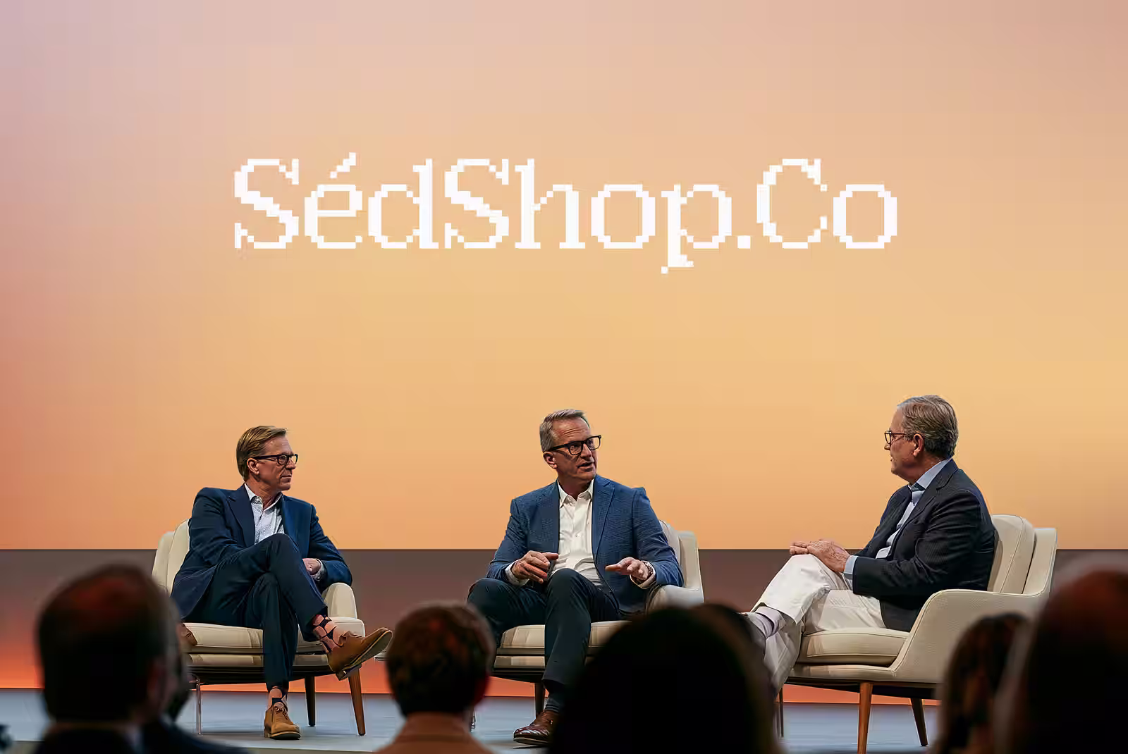A wide conference stage screen showing the SedShop.Co design behind three panel speakers at dusk.