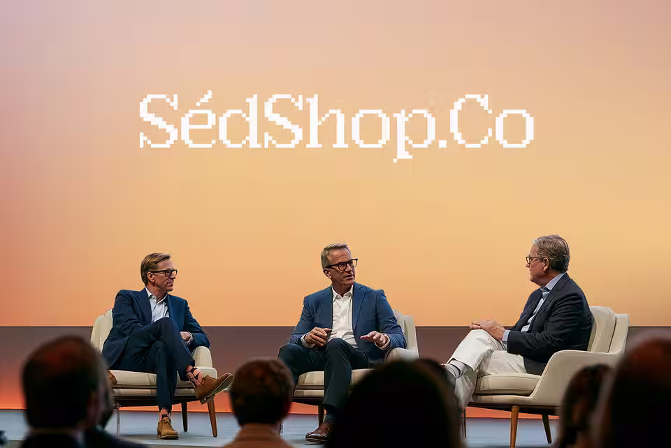 A wide conference stage screen showing the SedShop.Co design behind three panel speakers at dusk.