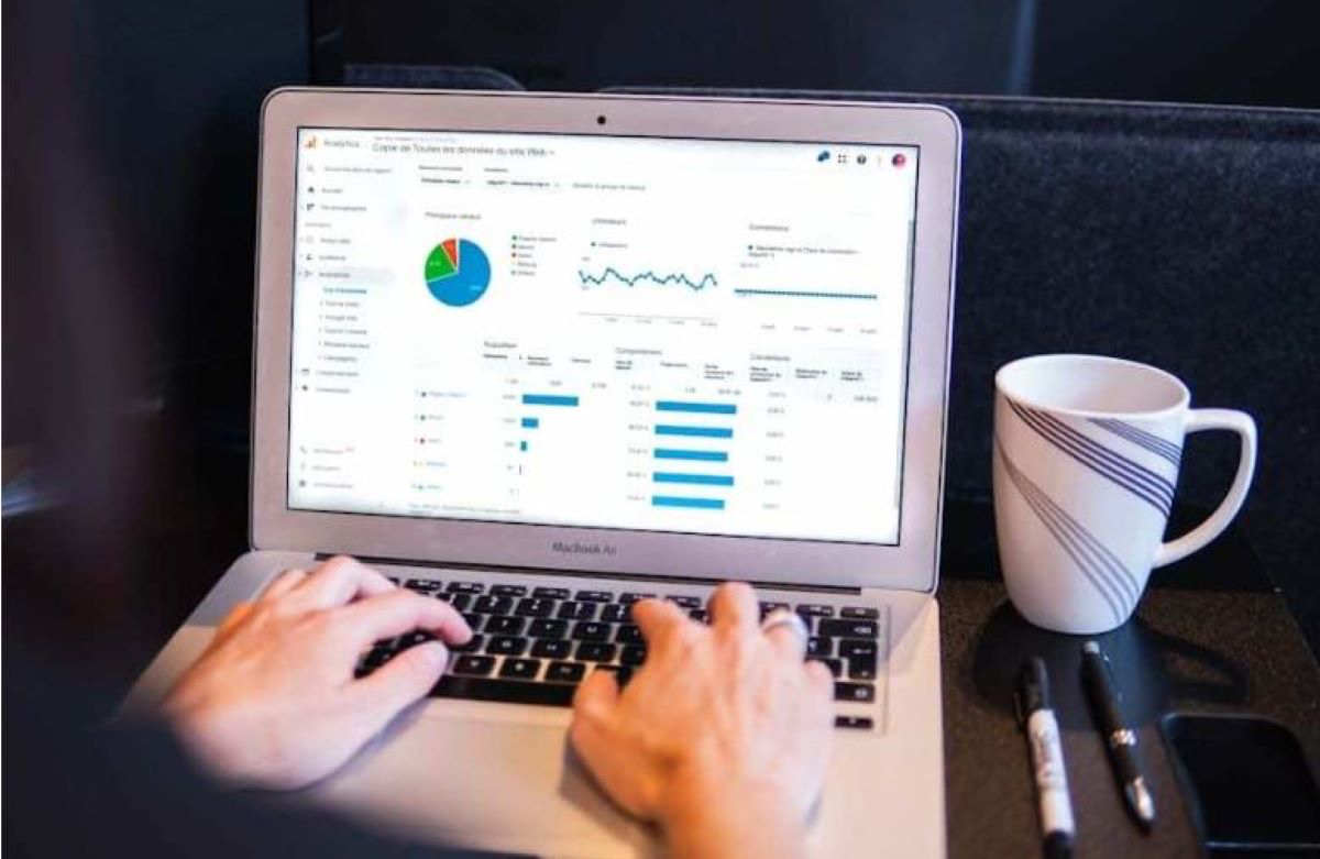 Image of someone preparing to type on a laptop that is sitting on a table or desk. The laptop has two graph and a chart displayed on the screen. There is also a white coffee mug sitting beside the laptop. 