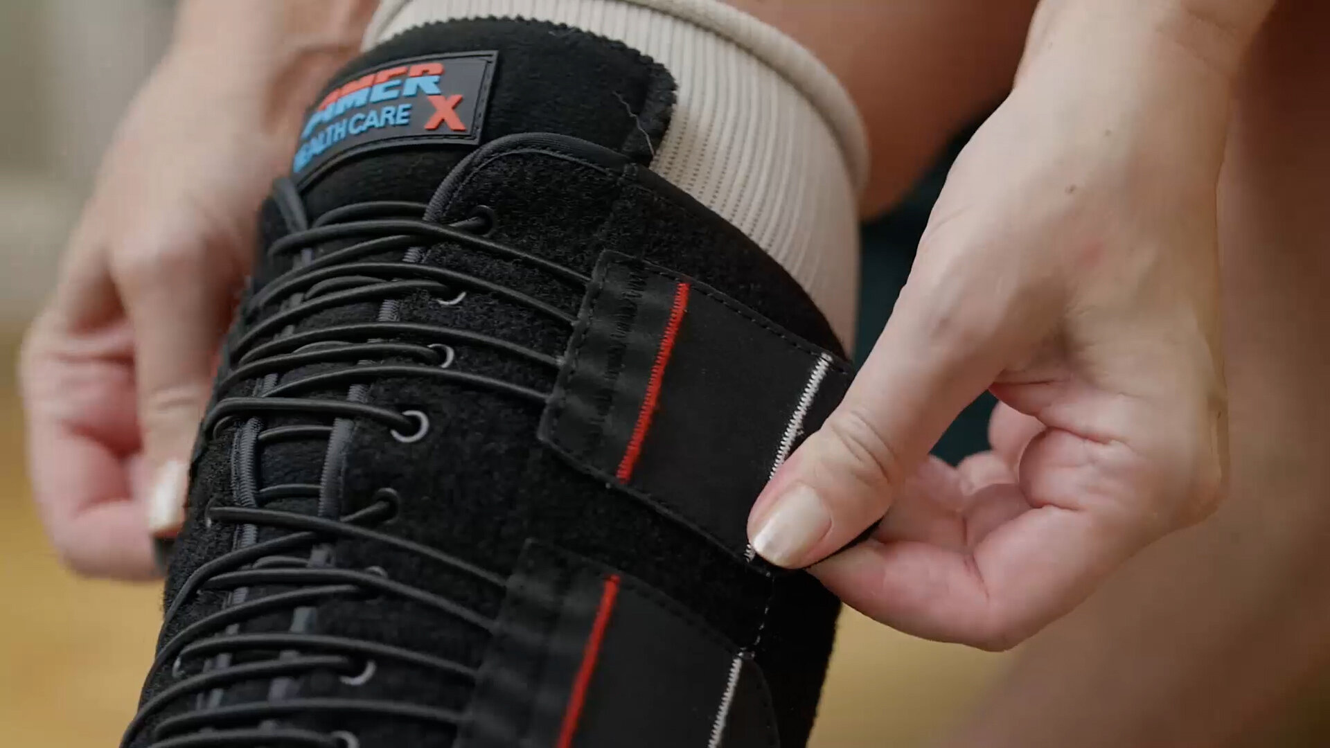 A close-up shot shows a person adjusting the straps of a black knee brace. The brace has "AMER HEALTHCARE X" written in white and red on its upper part. The person's leg is covered by a light-colored sock beneath the brace.