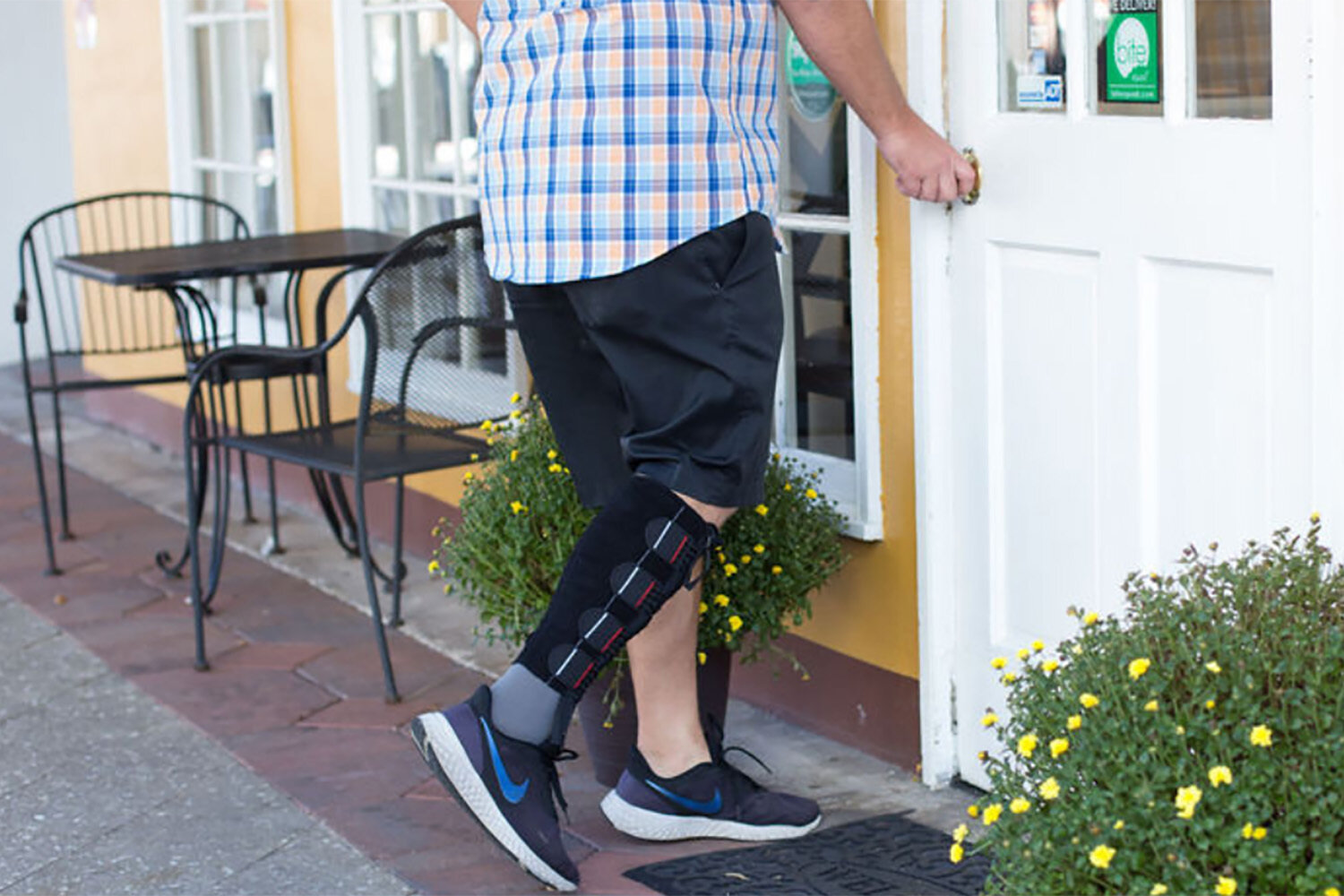 A person wearing black shorts, a blue and orange plaid shirt, and athletic shoes with a black compression sleeve on their lower left leg, opens a white door to a building. There are two outdoor metal tables with chairs outside the building, and a green plant with yellow flowers by the door.
