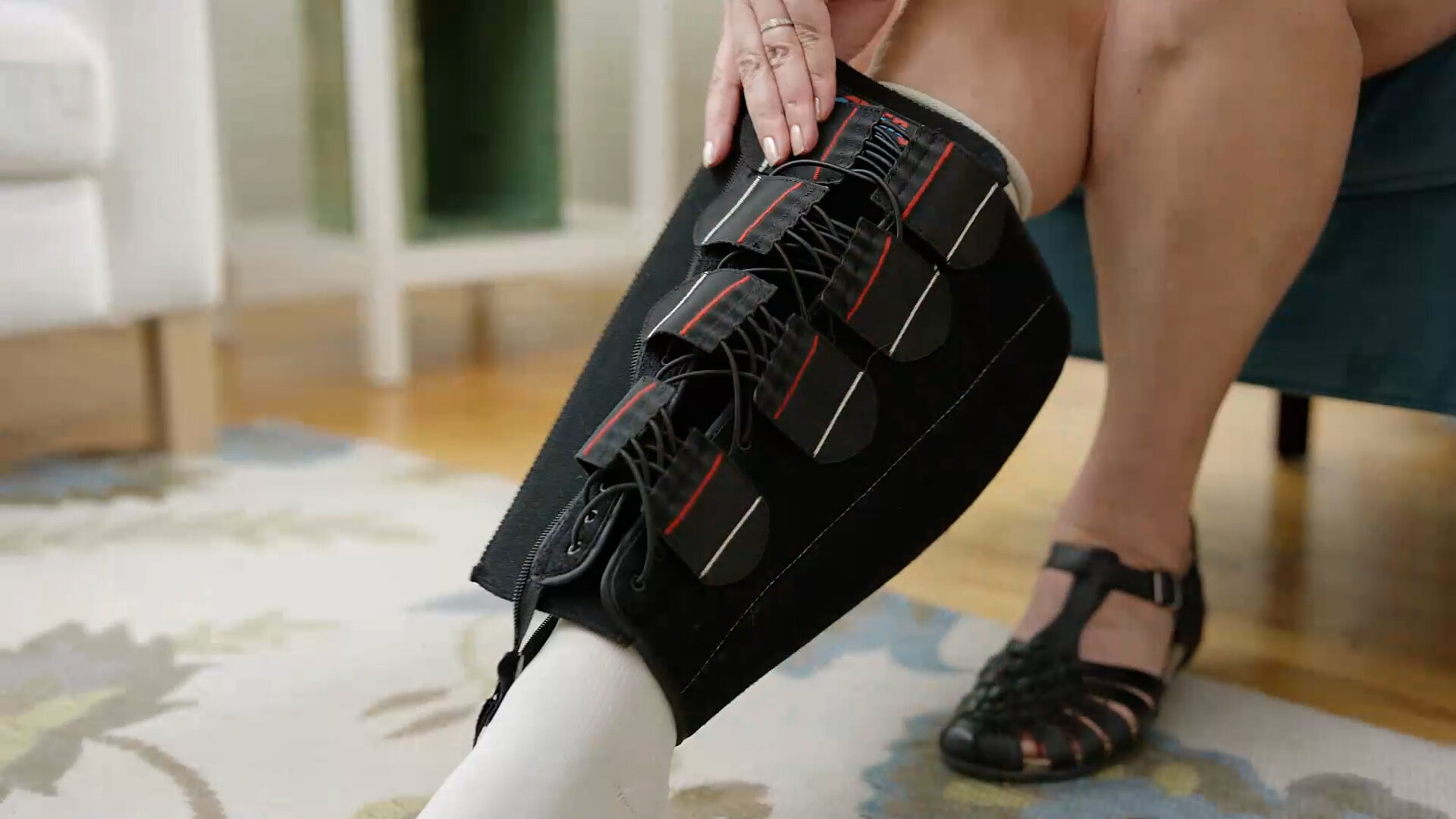 A person is adjusting a black compression device on their lower leg. The device has multiple straps with red and black accents. The person is wearing a white sock and black sandals.