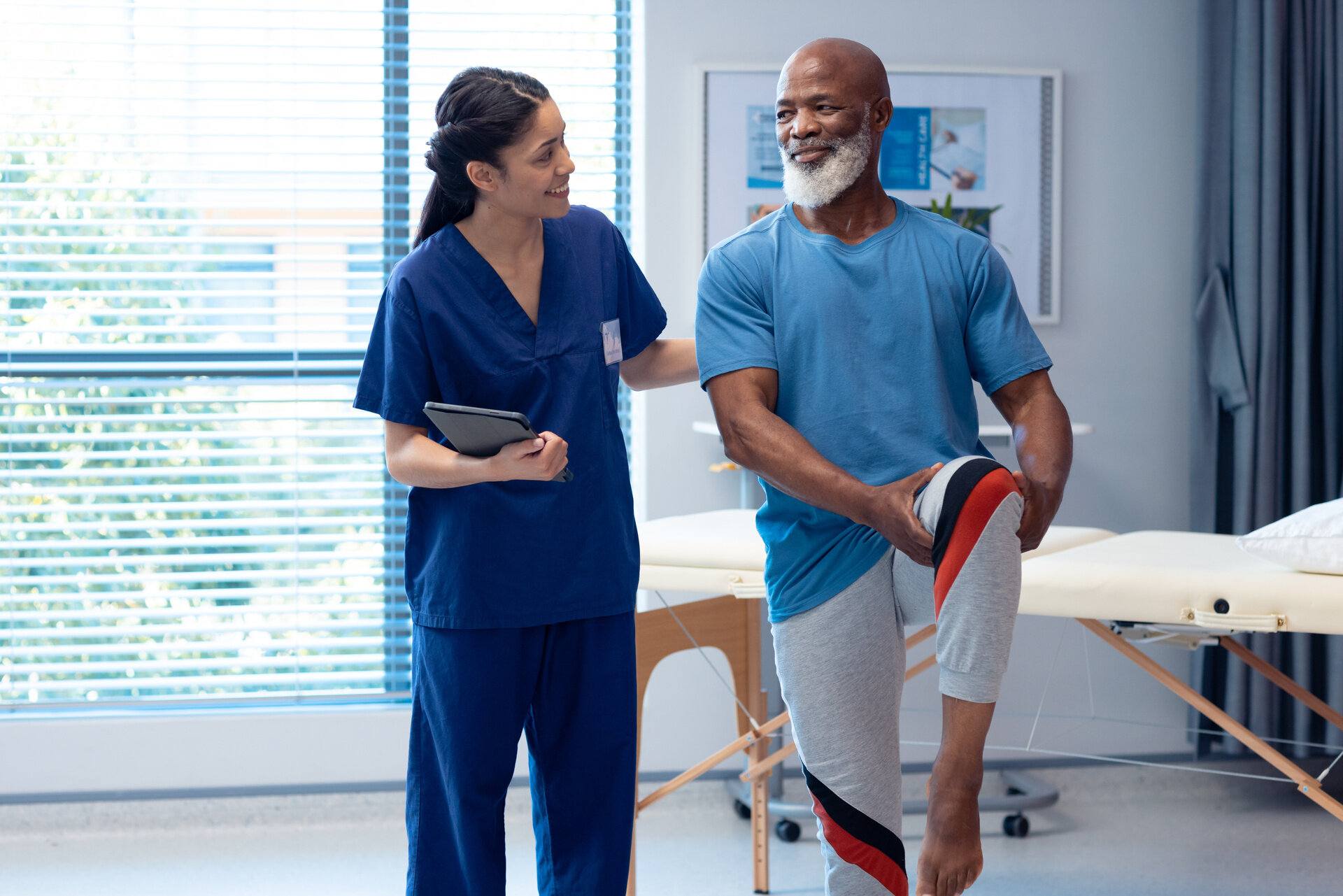 A physical therapist, dressed in blue scrubs, assists an older Black man with a leg exercise. The man is wearing a blue t-shirt and grey sweatpants with red and black stripes on the side. He is holding his right knee with both hands while standing on his left leg. The therapist is smiling at him, and they appear to be in a clinic or gym.