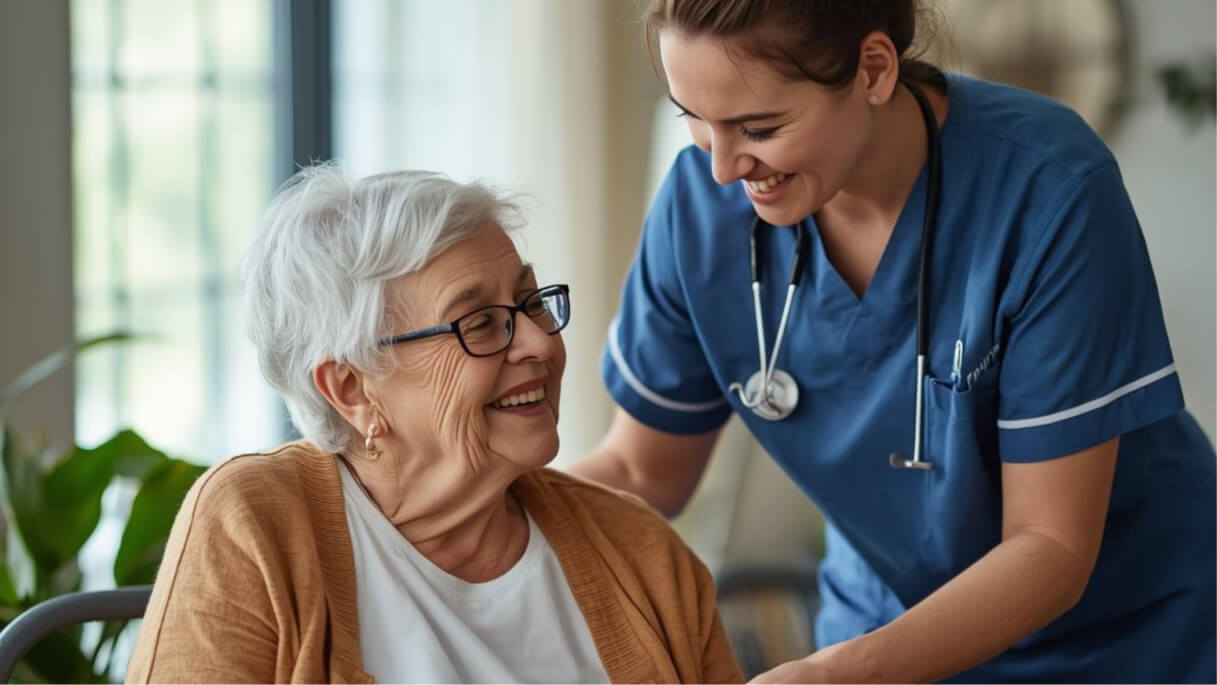 Smiling nurse in blue scrubs attending to a happy elderly woman with white hair and glasses.
