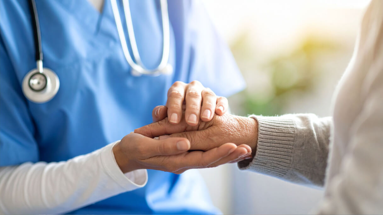 Healthcare professional in blue scrubs gently holding an elderly person's hand.