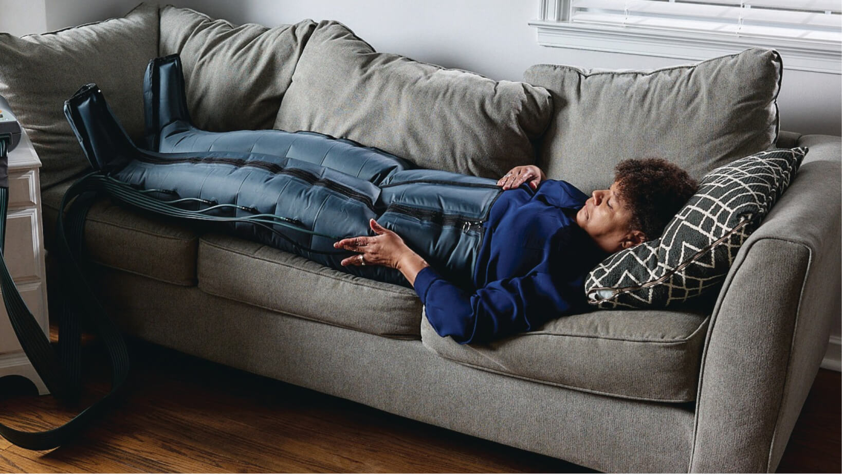 Woman lying on a couch wearing a full-body compression therapy suit connected to a device.