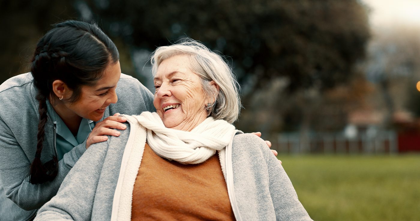 Young woman smiling and leaning towards an elderly woman sitting outdoors in a park, both appearing happy.