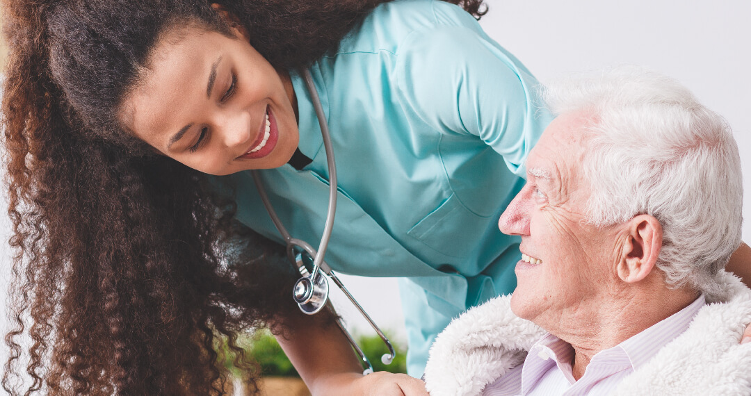 Smiling female healthcare professional with a stethoscope leaning toward an elderly man who is sitting and smiling back.