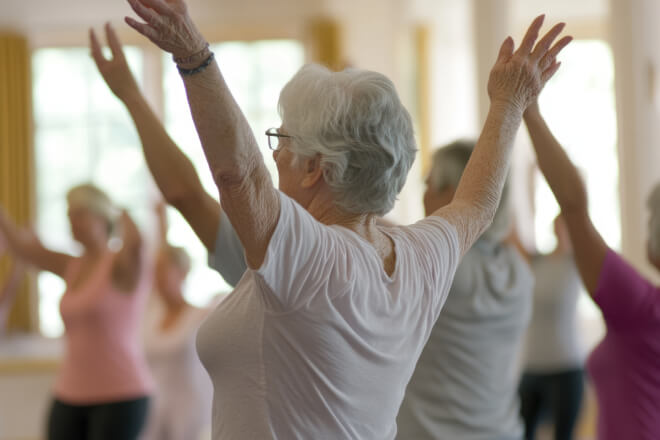 Group of elderly people exercising with arms raised in a bright room.