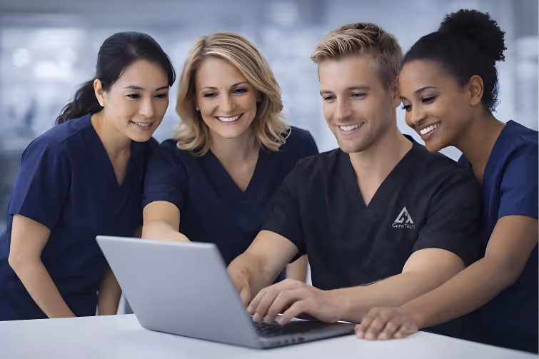 Four medical professionals in scrubs gathered around a laptop, smiling and collaborating.