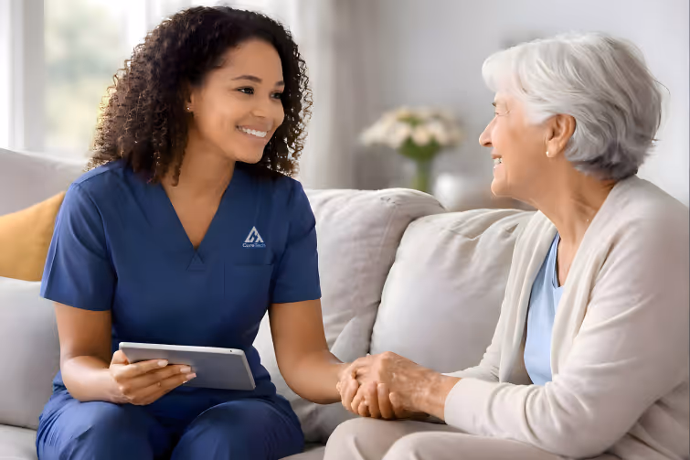 Smiling nurse in blue scrubs holding a tablet while sitting and holding hands with an elderly woman on a couch.