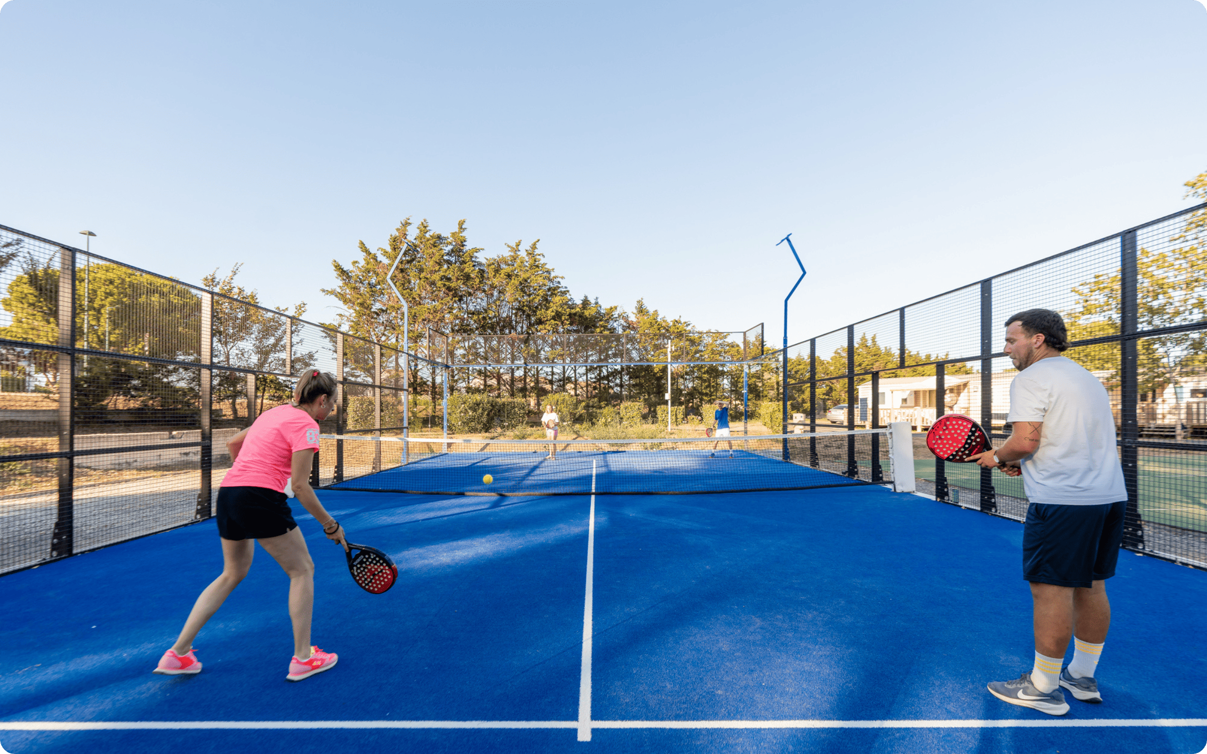 Outdoor recreational area with a blue paddle tennis court, playground with slide and climbing equipment, picnic tables, and a fitness zone with exercise stations.
