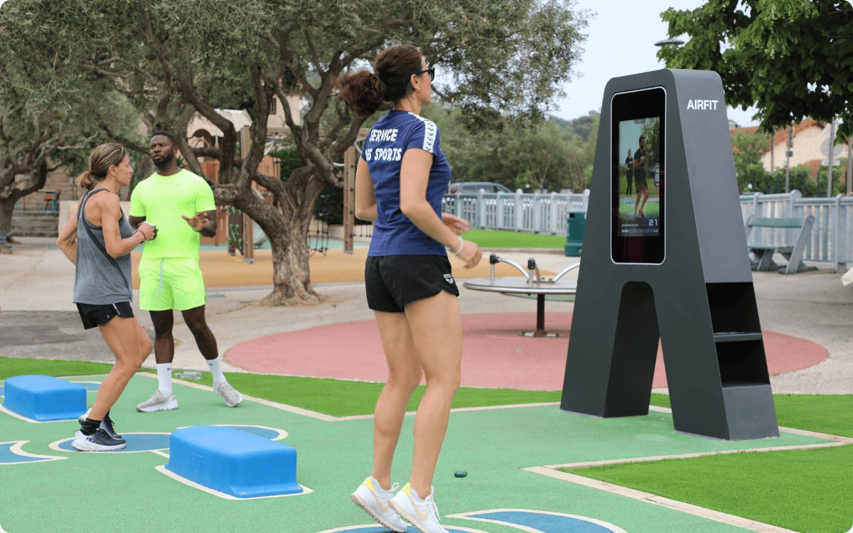 Outdoor recreational area with a blue paddle tennis court, playground with slide and climbing equipment, picnic tables, and a fitness zone with exercise stations.