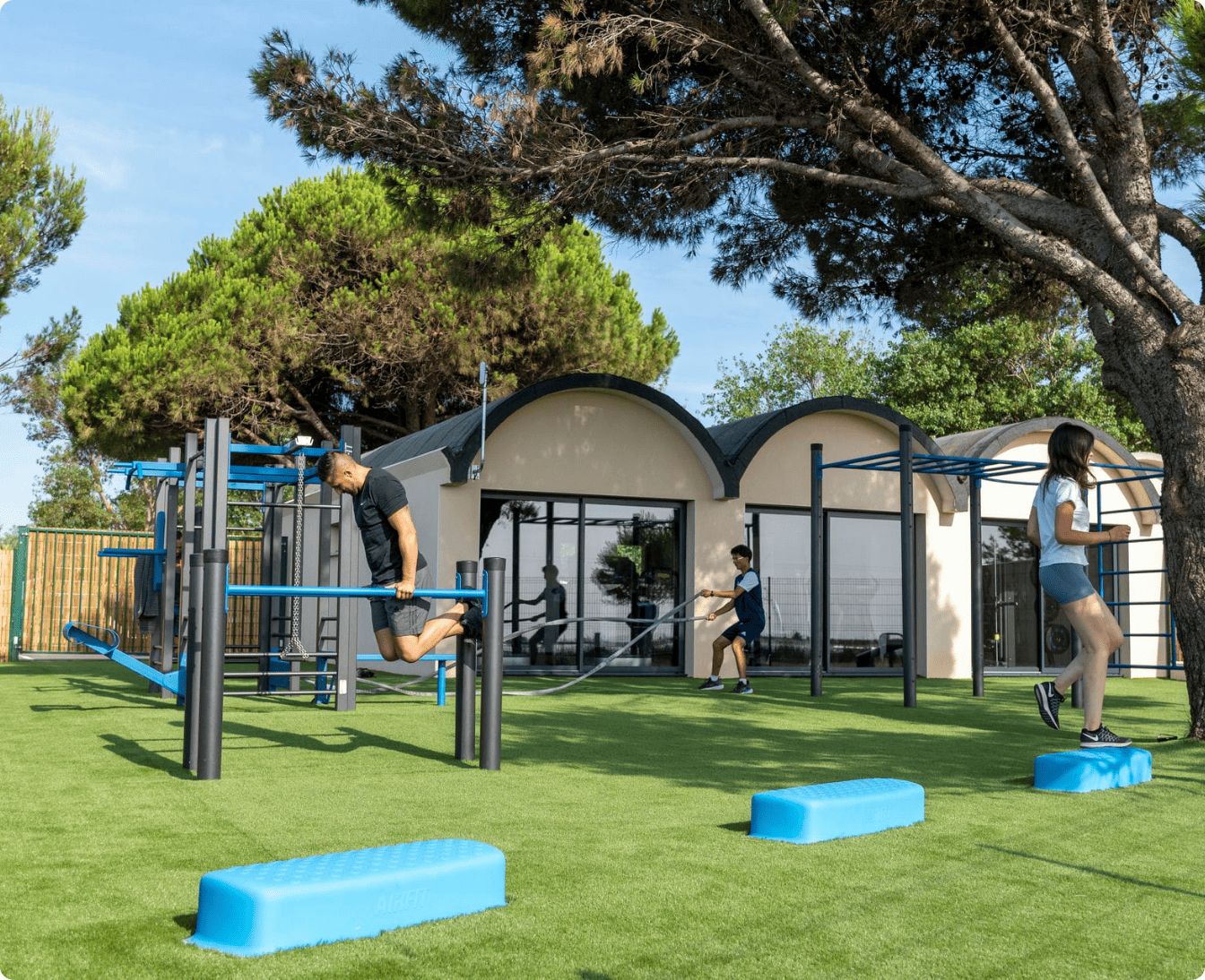 Outdoor recreational area with a blue tennis court, picnic tables on grass, a playground with slide, and a fitness zone with exercise equipment.