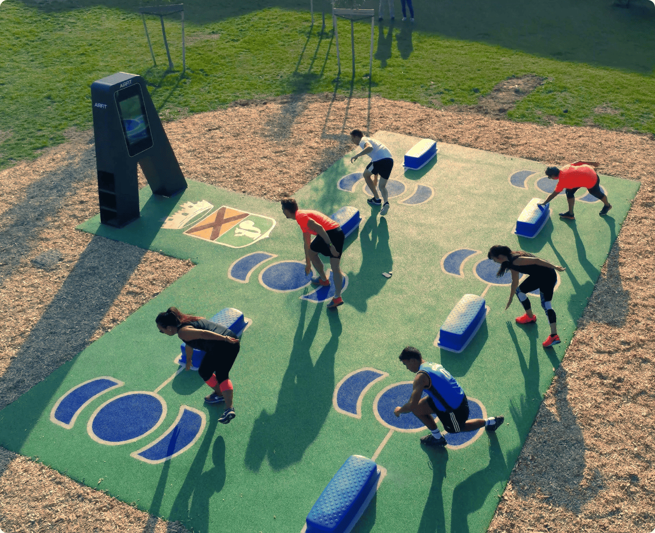 Outdoor fitness park with various workout equipment on green artificial turf near residential buildings under a partly cloudy sky.
