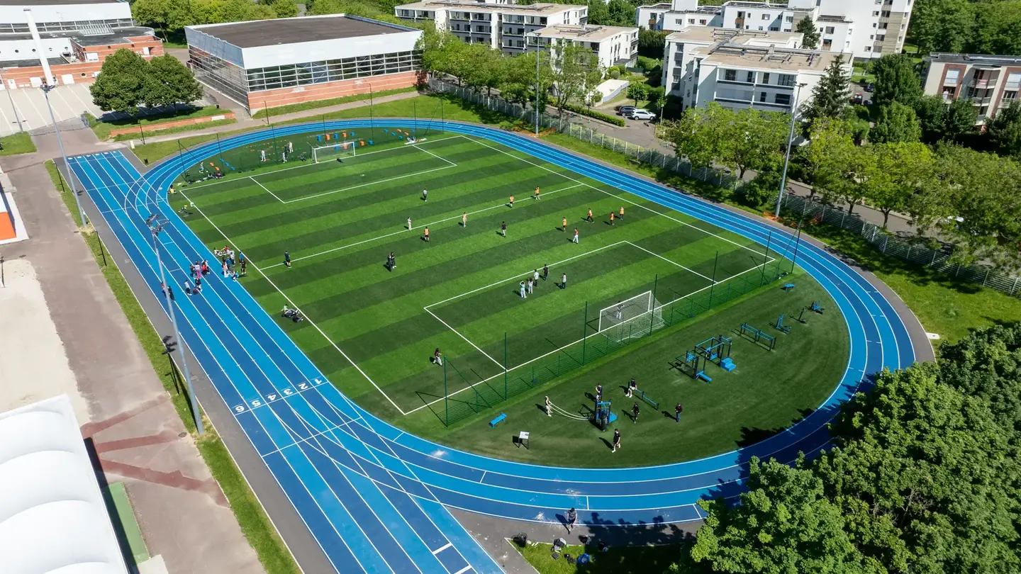 Vue aérienne du stade Pierre de Coubertin à Montigny-le-Bretonneux : la demi-lune de cross-training au premier plan, le terrain de football au centre, la demi-lune de motricité au fond, dans les virages de la piste d'athlétisme