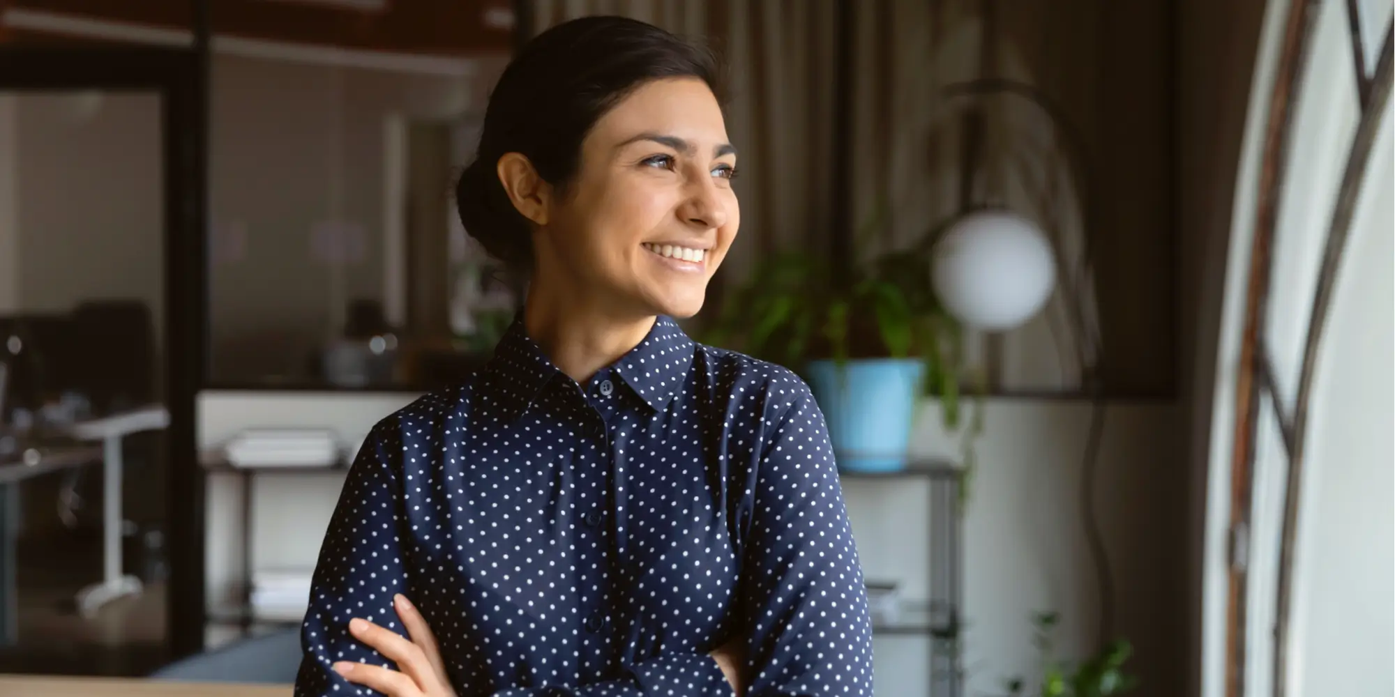 Smiling woman in a blue polka-dot shirt standing with arms crossed in a modern office space.