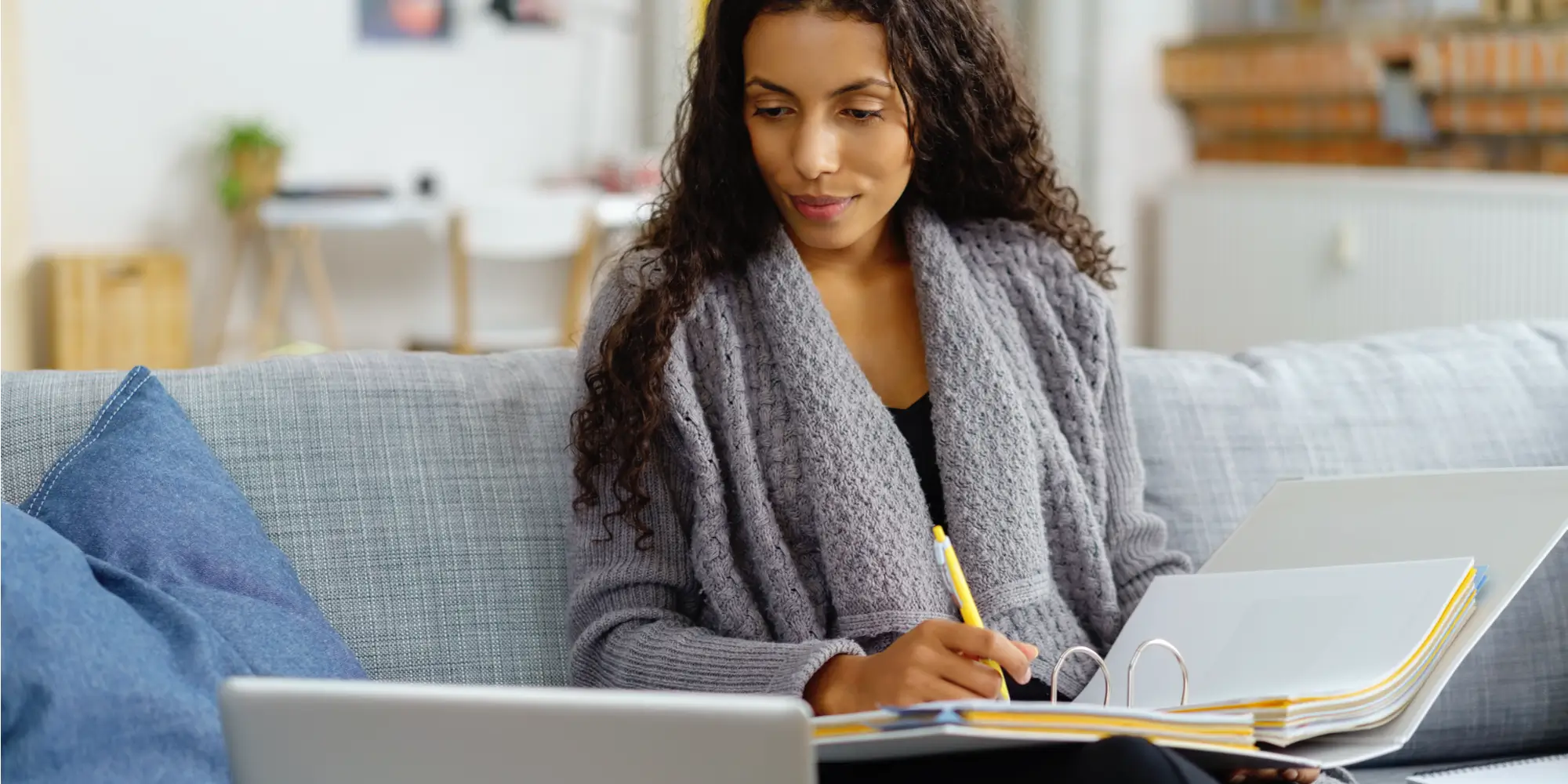 Young woman sitting on a couch writing with a pen in a binder while looking at a laptop.