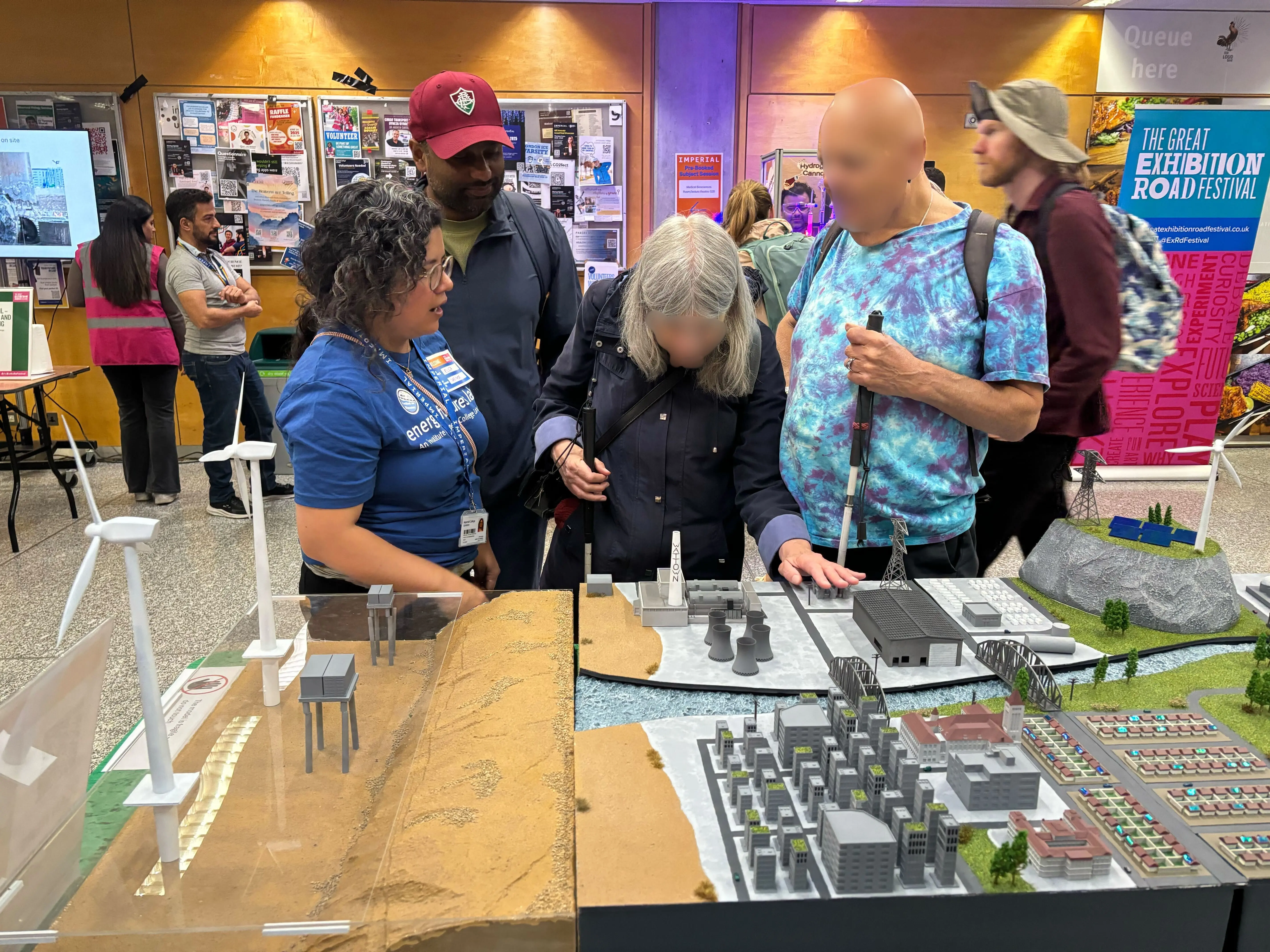 A visually impaired woman, guided by a volunteer, explores a tactile model of an energy landscape featuring wind turbines, buildings, and power stations. Another VIP stands beside her using a white cane. An exhibition staff member in a blue T-shirt explains the model, while other attendees observe in the background. A banner for the Great Exhibition Road Festival is visible behind them.