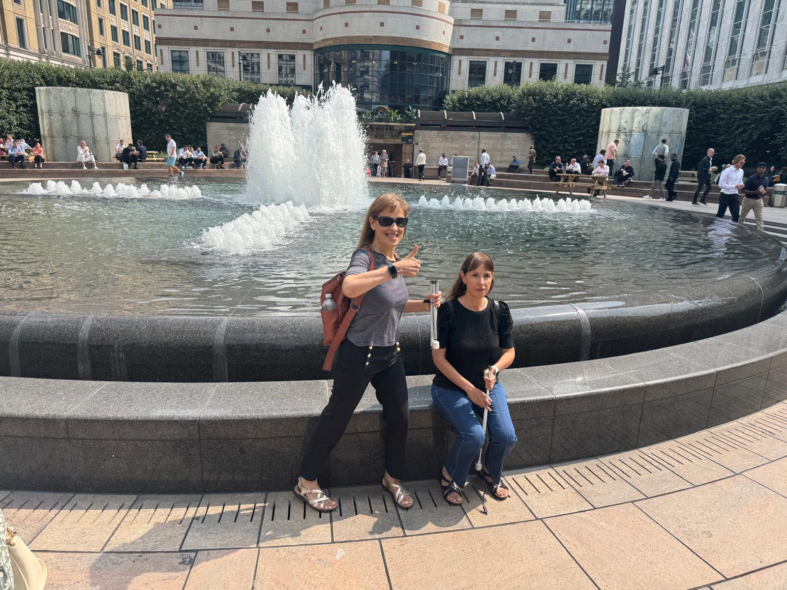 VIP Fabiana posing with a thumbs up and VIP Paula smiling facing the camera. They are in front of a fountain