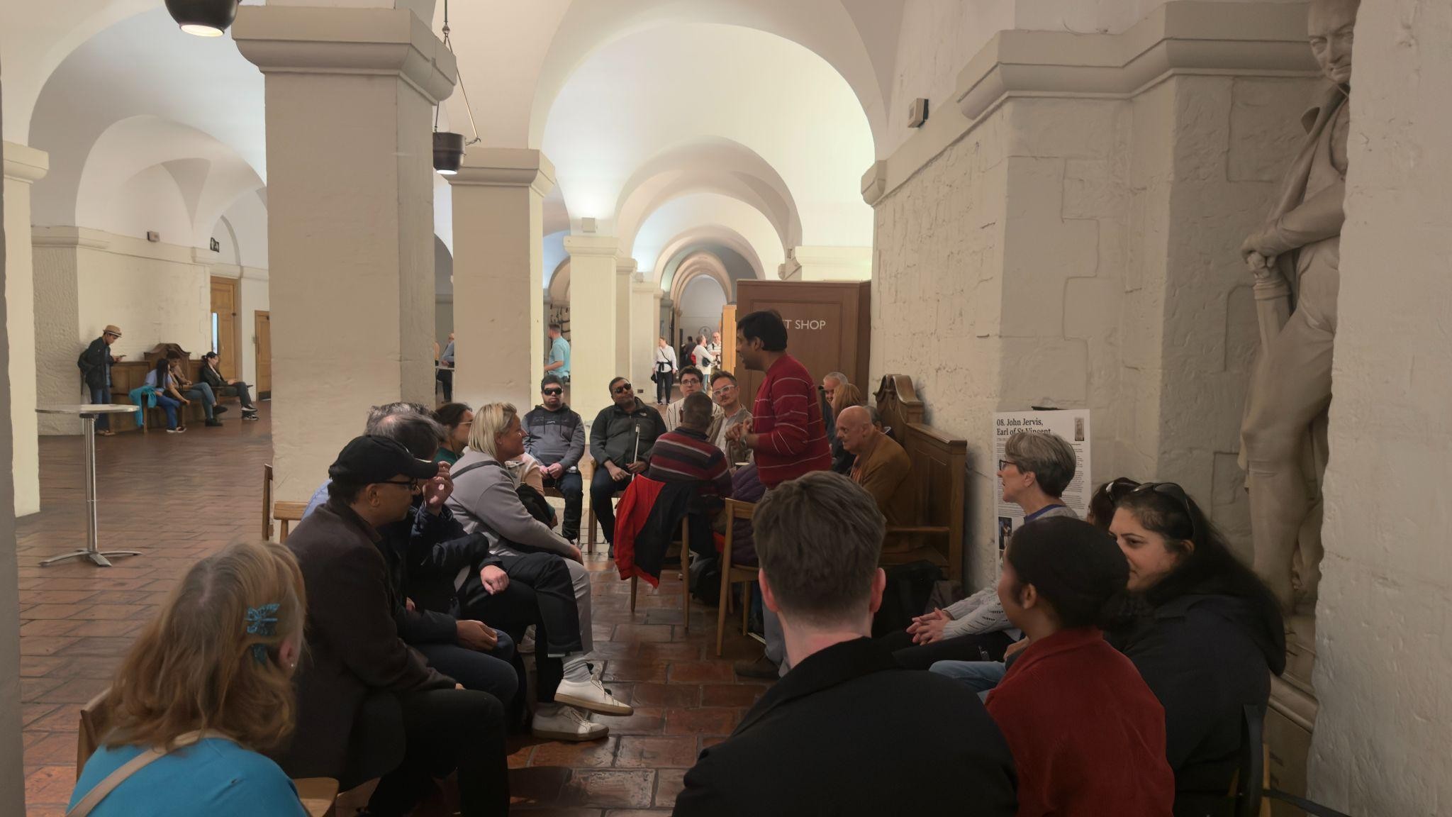 A group of people seated in a semi-circle inside a historic building with arched ceilings, participating in a Sainsbury’s volunteer event. One person is standing and speaking to the group, while others listen attentively. The setting appears warm and collaborative, with statues and stone walls in the background.