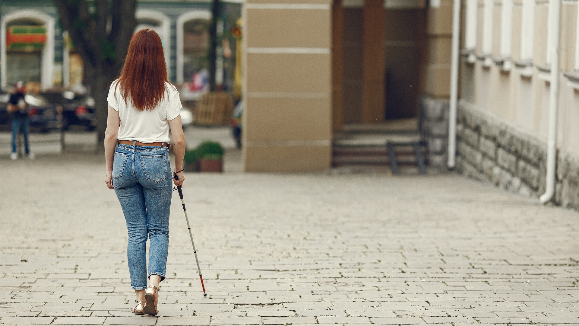 A visually impaired person walking independently outdoors using a white cane on a paved street.