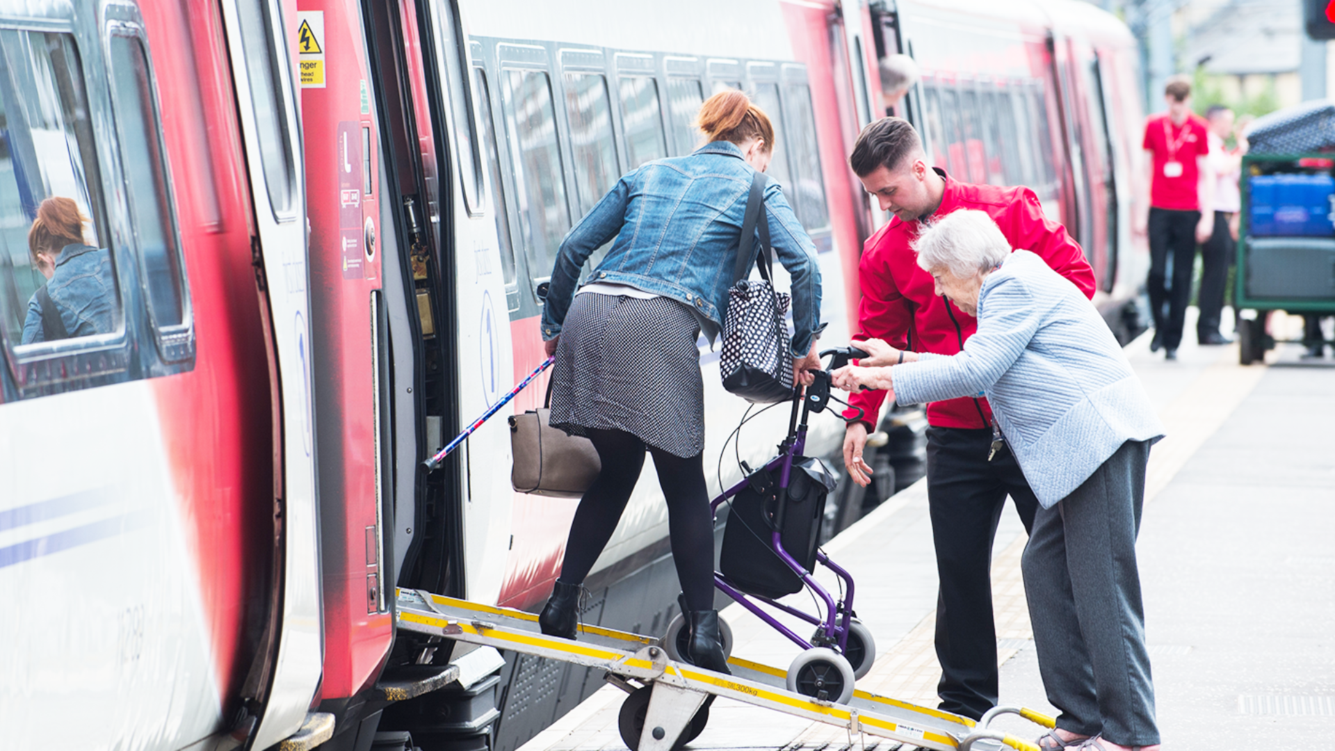 An older person being assisted by railway staff while boarding a train using a ramp at a UK station.