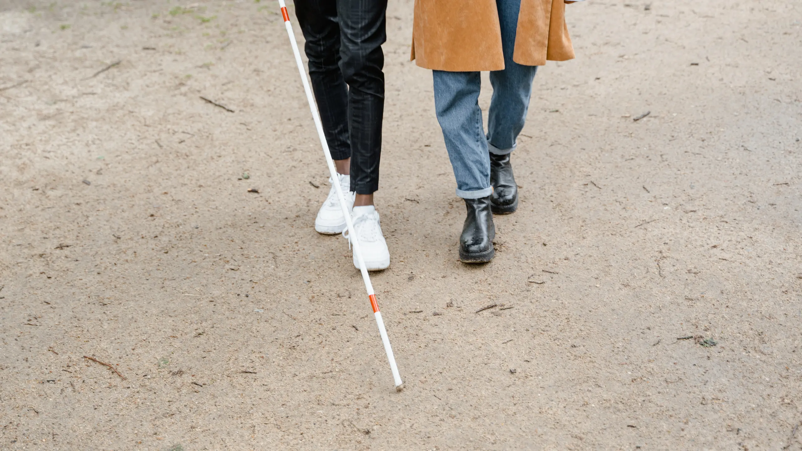 Visually impaired person walking alongside a companion while using a white cane on an outdoor path