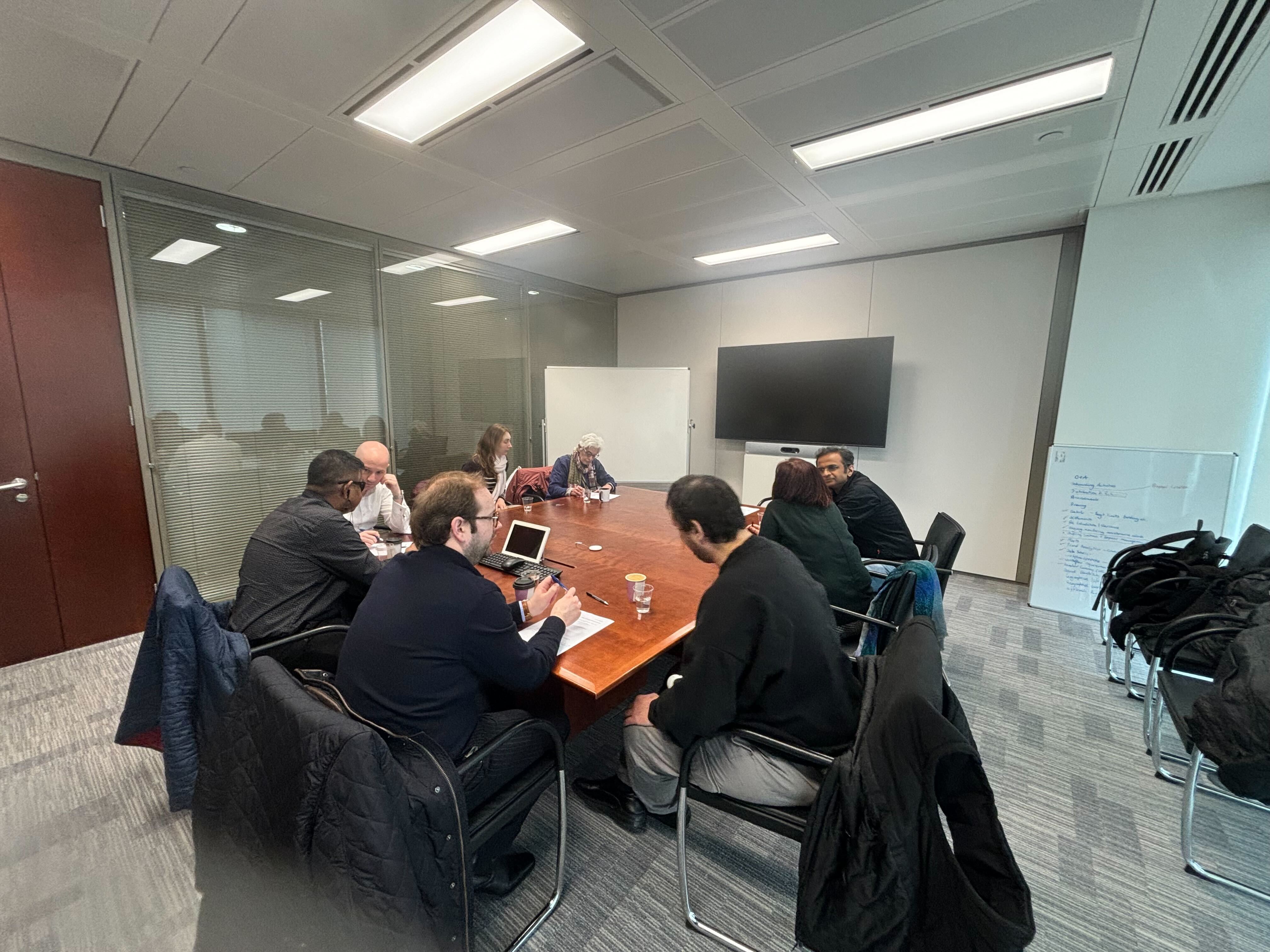 A group of corporate volunteers seated around a large wooden meeting table in a modern office conference room during the JPMC Corporate Volunteer Event. Participants are engaged in discussion, with notebooks, laptops, coffee cups, and printed materials on the table. A whiteboard and large screen are visible at the front of the room, creating a collaborative workshop setting.