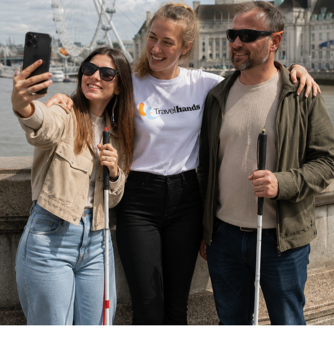 three people standing a travel hands volunteer posing for a selfies with young girl wearing tinted glasses who is holding her white cane and holding up her phone to take a selfie, on right there is a middle aged man smiling and holding his white mobility cane.