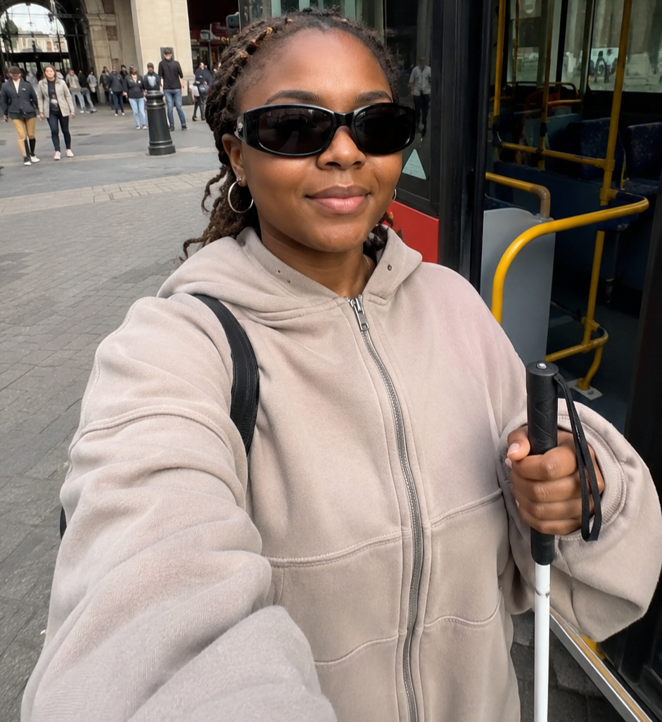 a blind woman making vlog with her phone, she is wearing a brown hoodie, dark tinted glasses and have braided hair and is holding her white mobility cane.