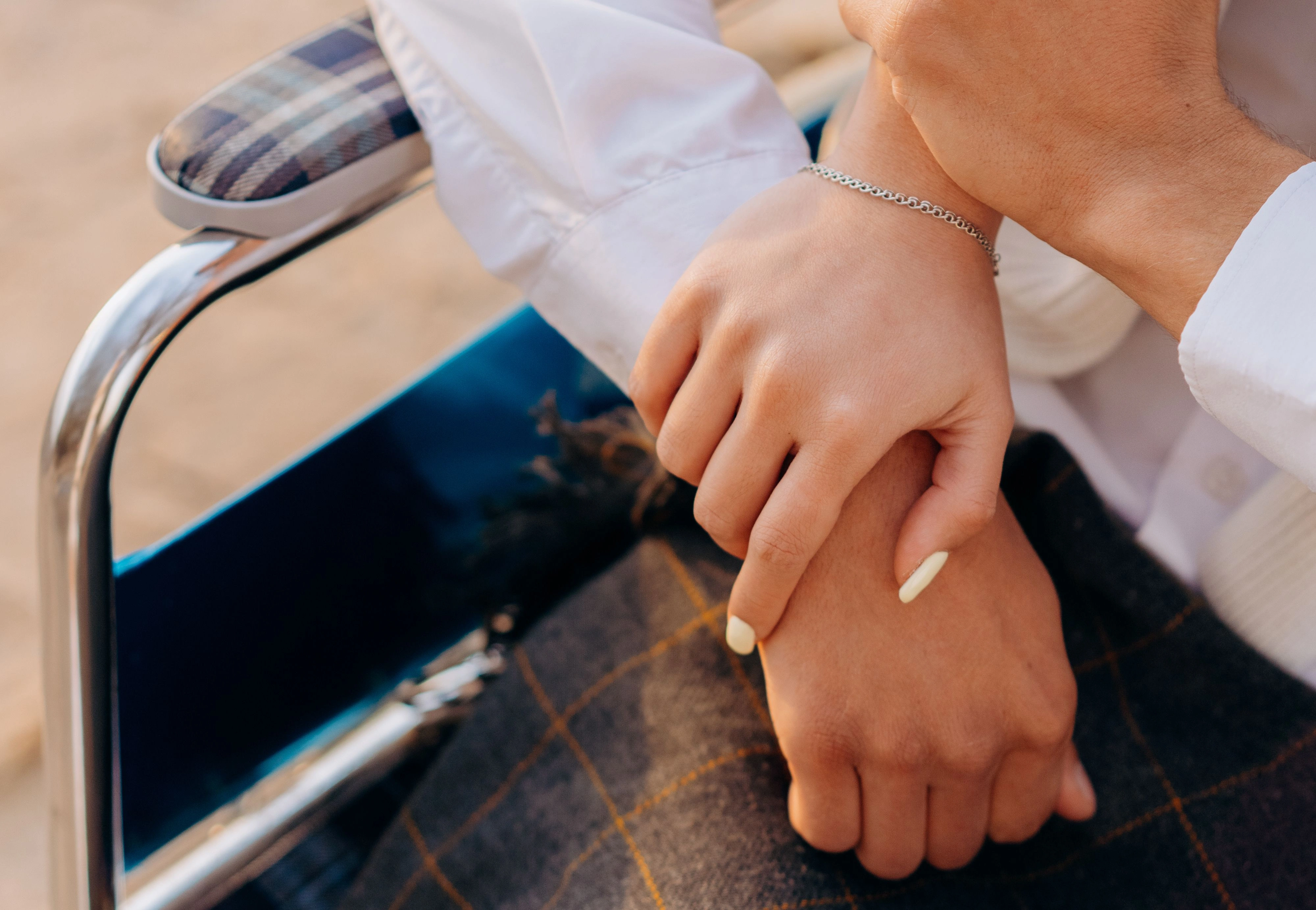 Close-up of one person's hand resting on the hand of someone seated in a wheelchair, a tartan blanket across their lap