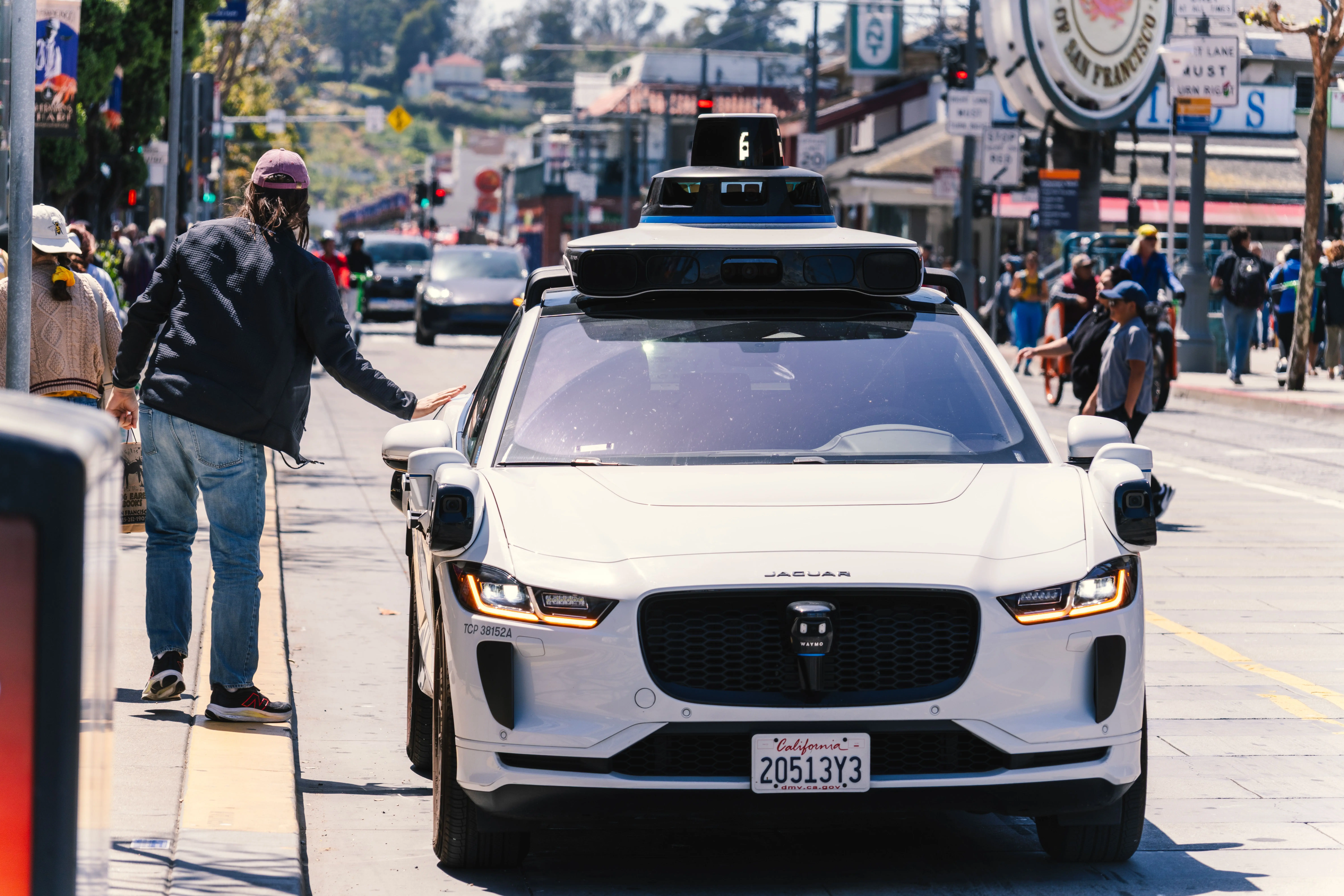 Self-driving Waymo car with roof-mounted sensors stopped in traffic on a busy San Francisco street as a pedestrian walks past with a hand on the bonnet