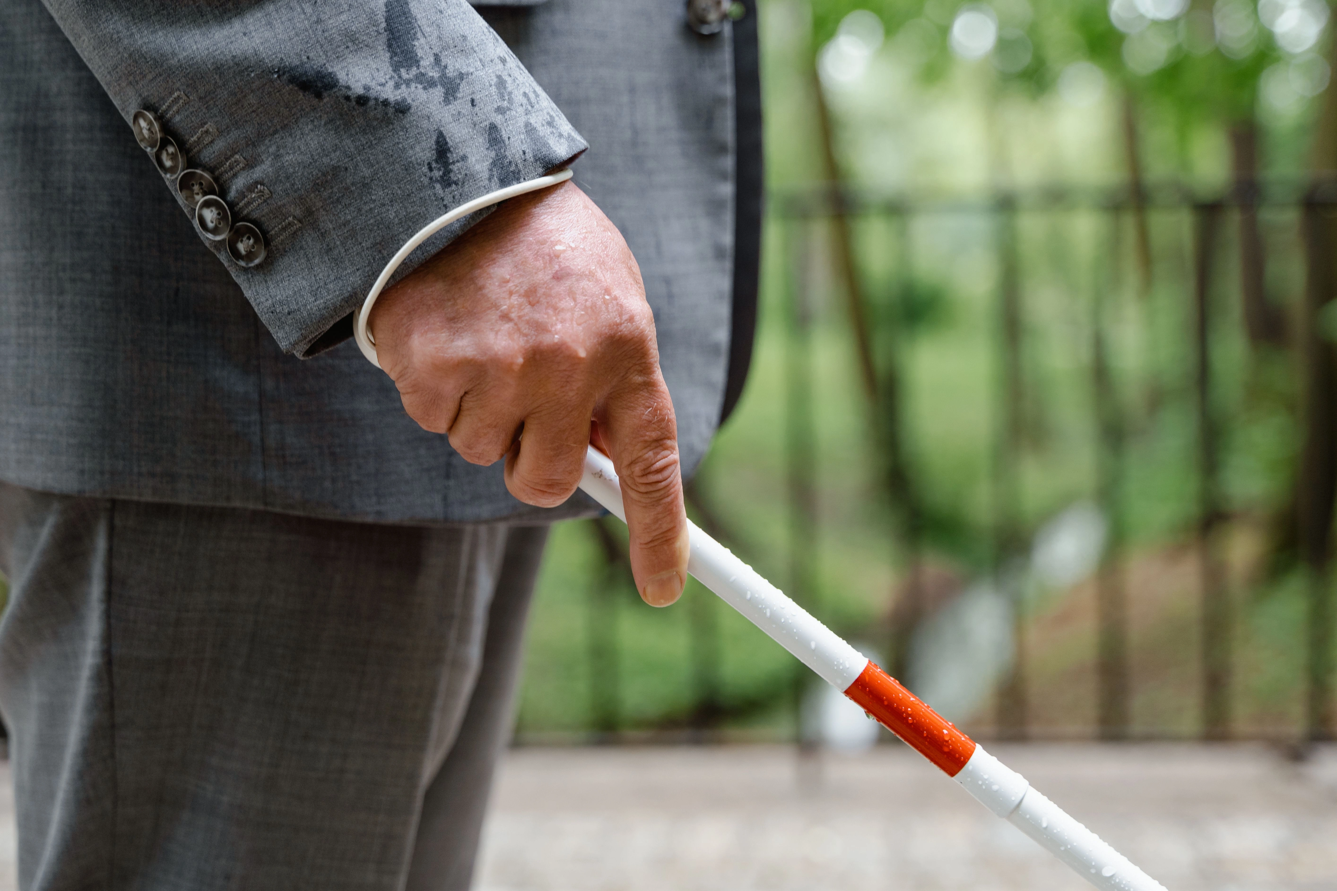 Close-up of a man's hand holding a white cane with a red band, rainwater beading on the cane and his sleeve