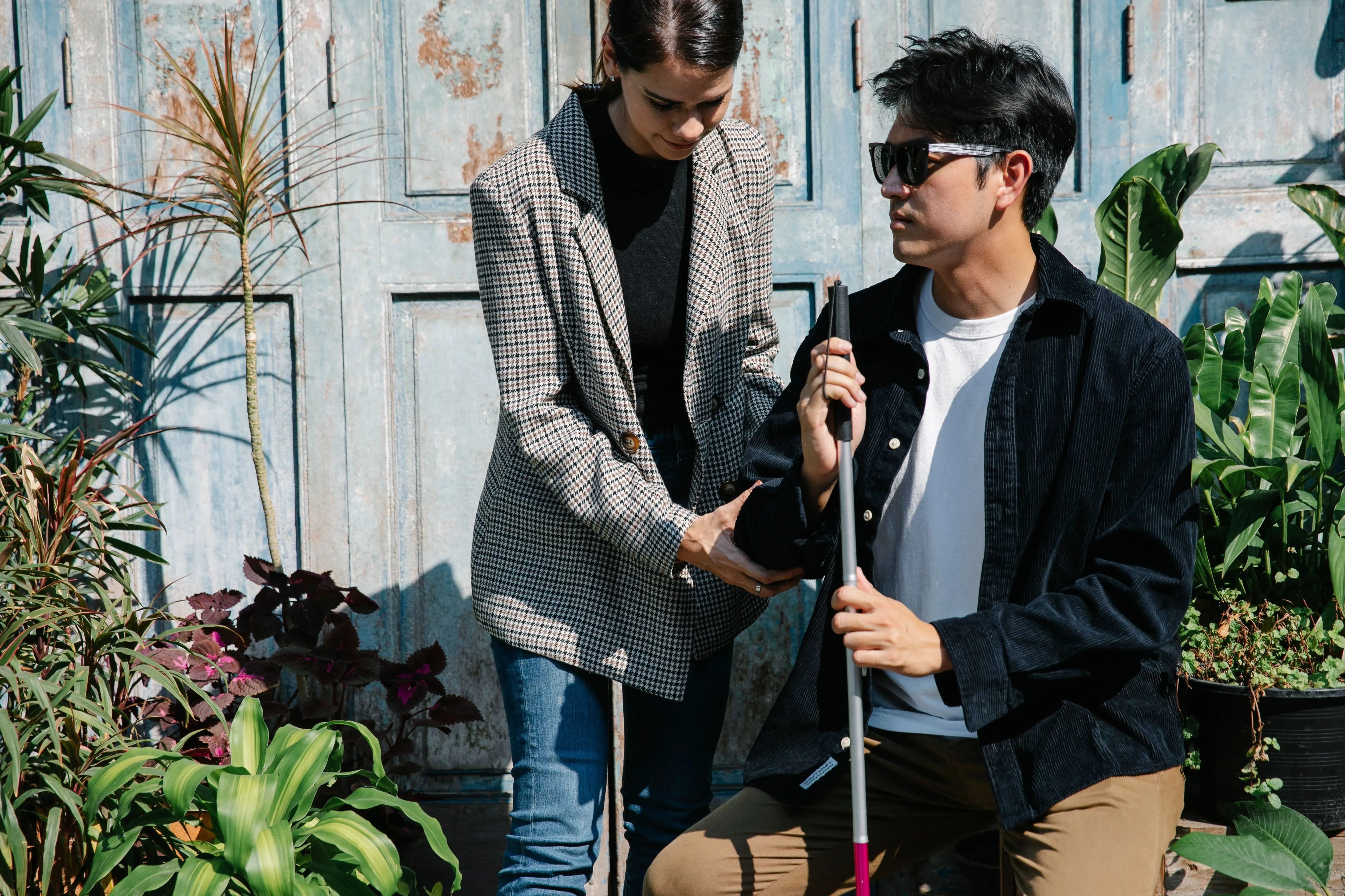 A woman guides a blind man's hand to his folded white cane as they sit outdoors beside a blue door surrounded by plants