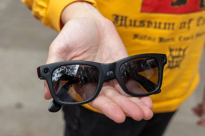 Black Ray-Ban Meta camera glasses resting in an open palm, with the small camera lens visible in the corner of the frame