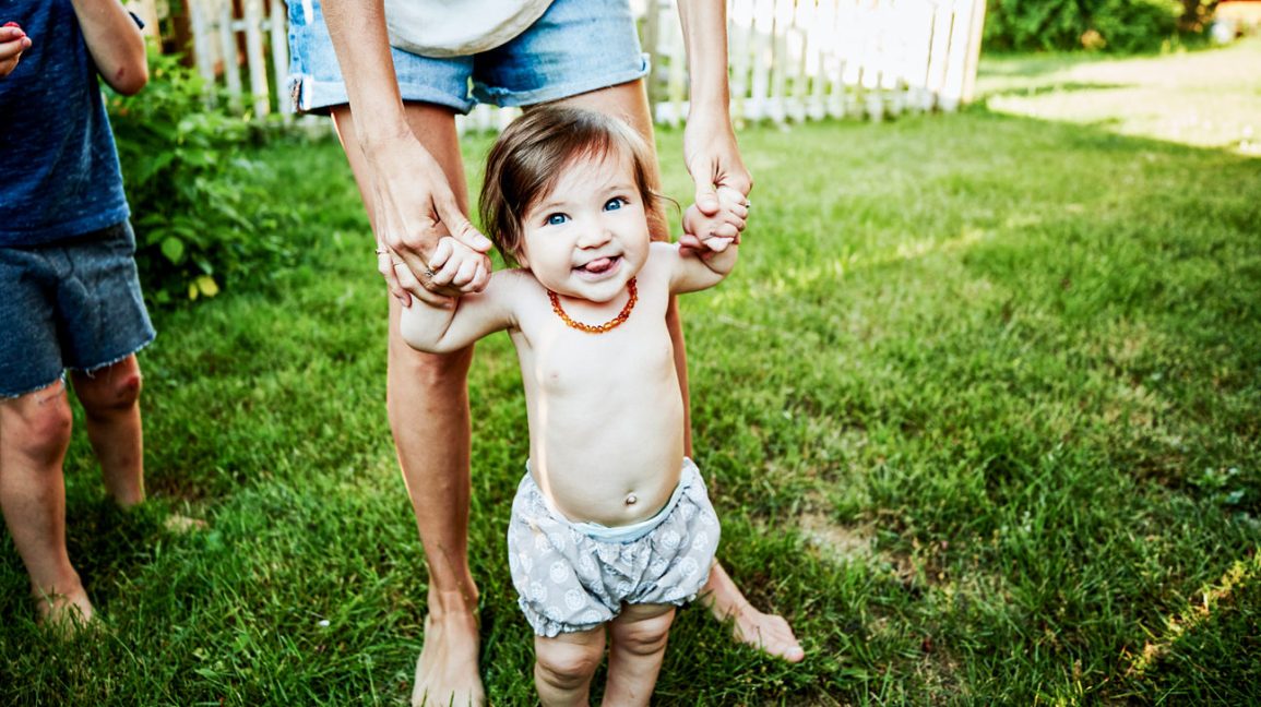 Mother-helping-infant-daughter-walk-in-backyard-on-summer-morning-1296x728-header-1296x728