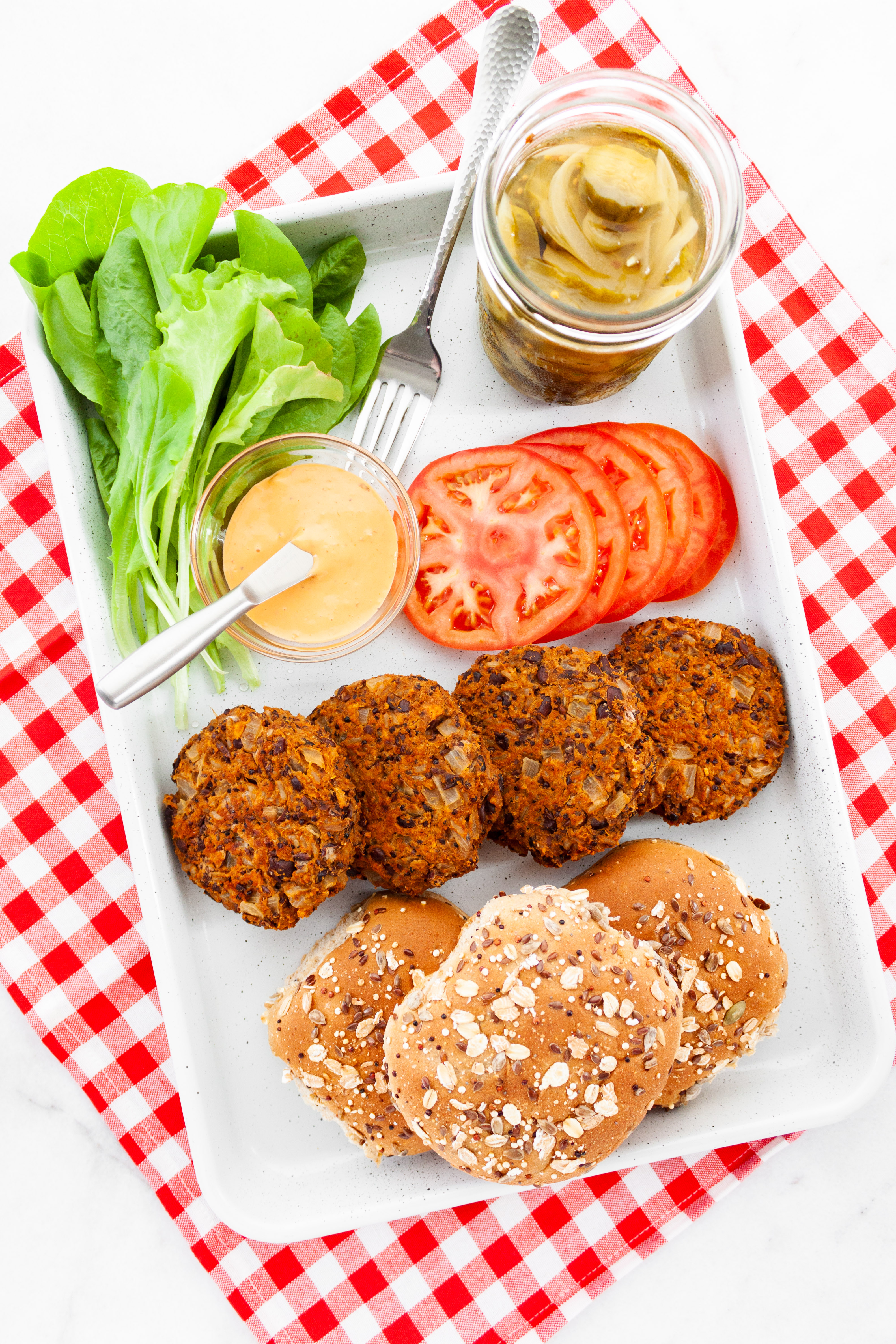 A white tray laid out with buns, burgers, sliced tomato, lettuce leaves, jar of pickles, and dish of burger sauce