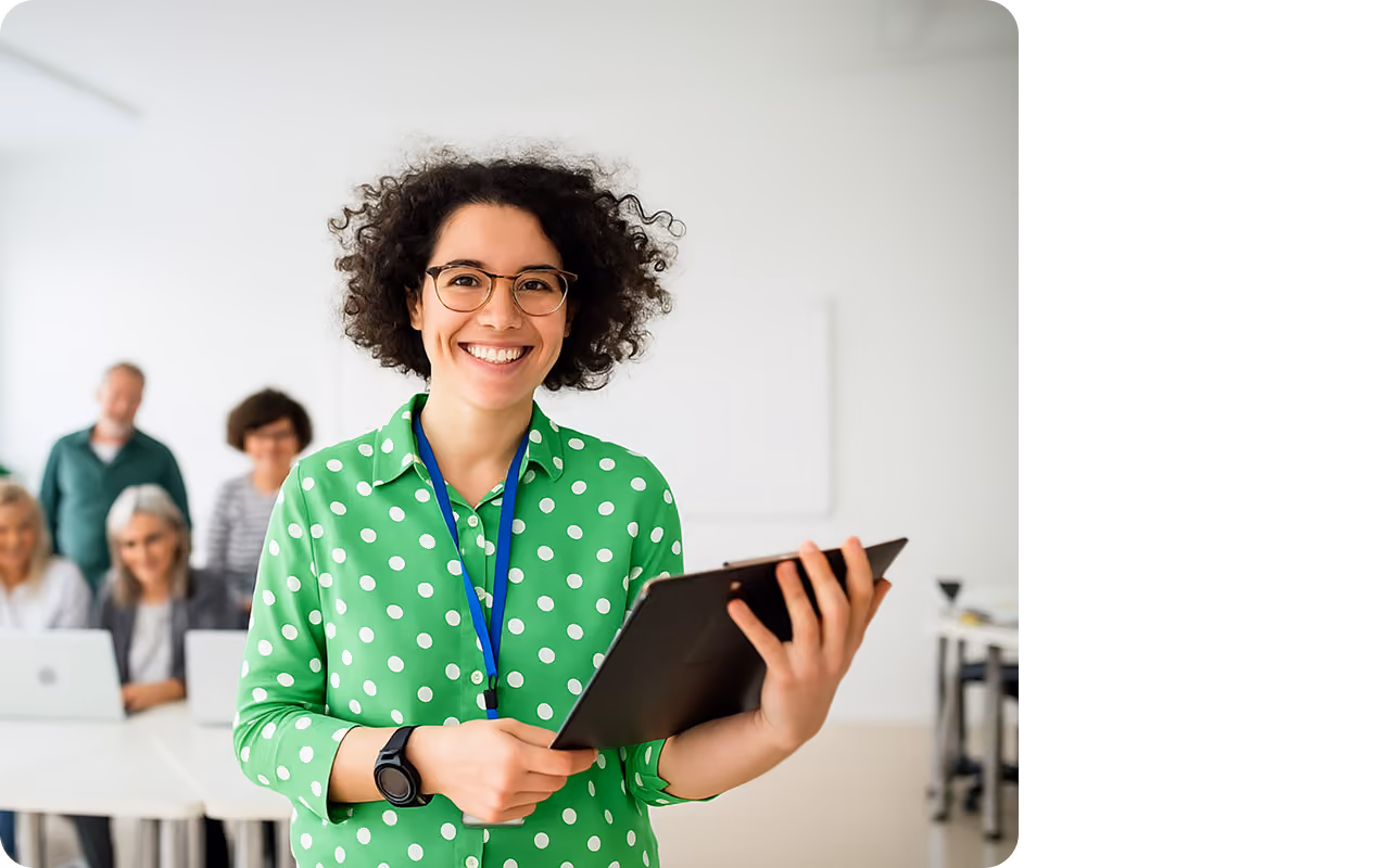 Educator holding a tablet and smiling in a classroom, with colleagues seated behind.