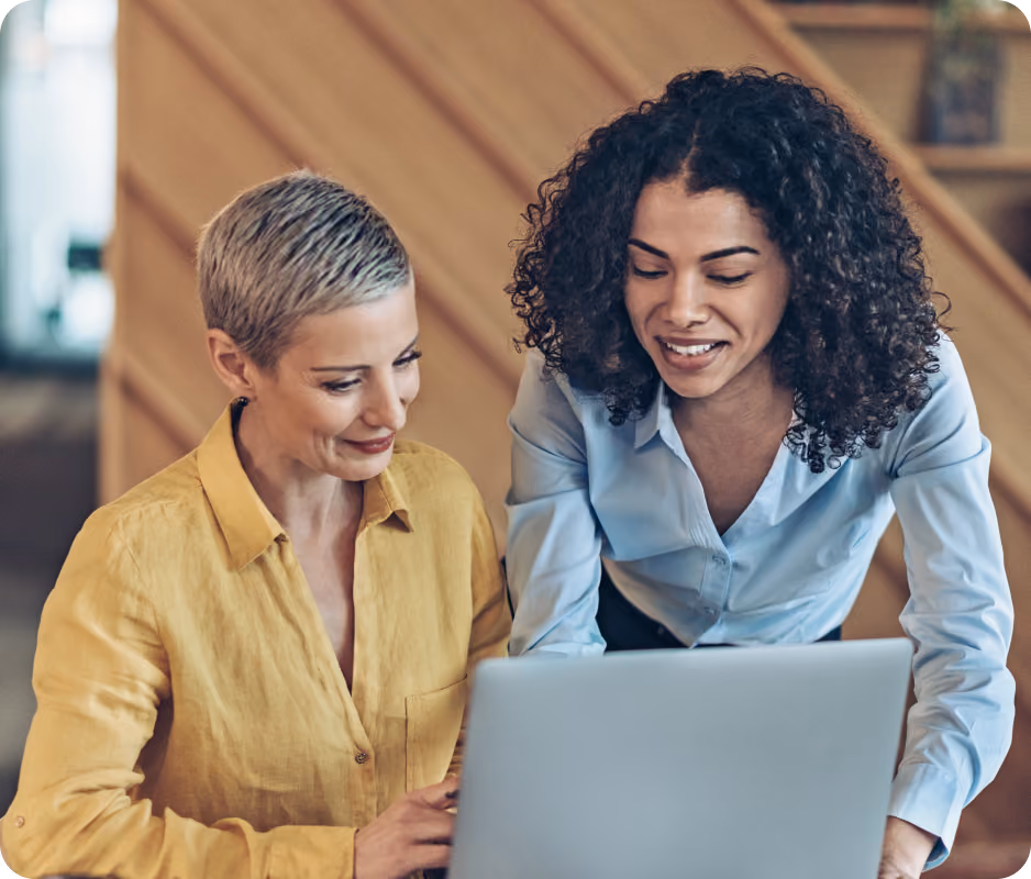 Two women looking at a laptop together, one seated in yellow shirt, other standing in blue shirt.