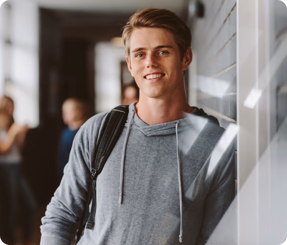 Smiling young man in a gray hoodie with backpack standing indoors near a window.