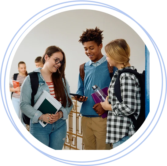 Three students with backpacks chatting and looking at a phone in a school hallway.
