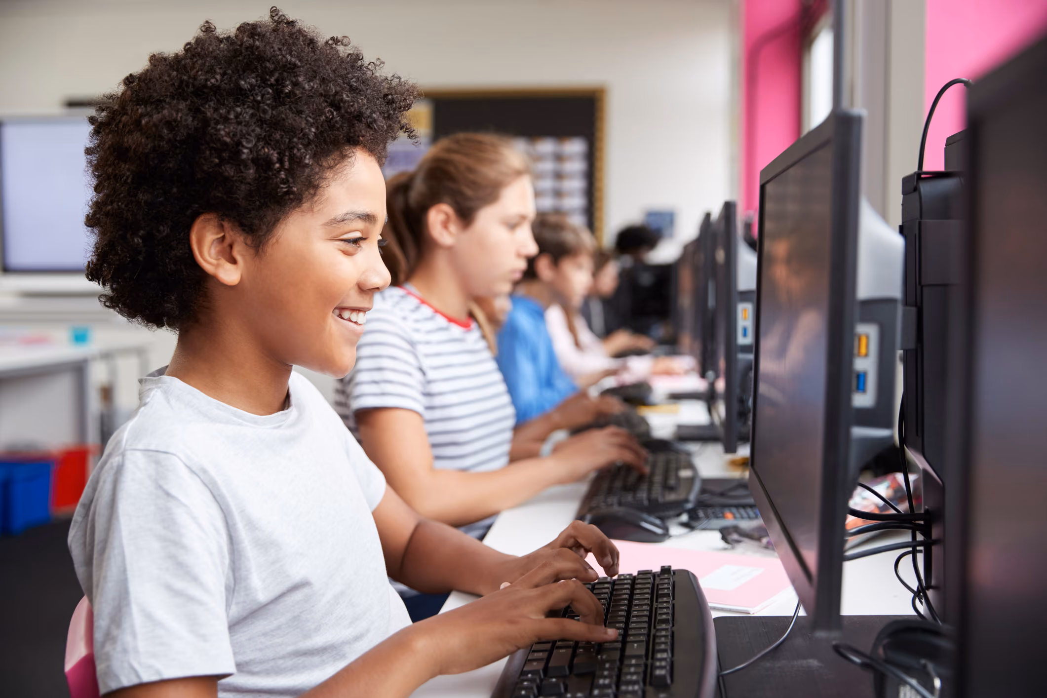 A row of children working on desktop computers in a classroom, with the closest child smiling while typing on a keyboard.